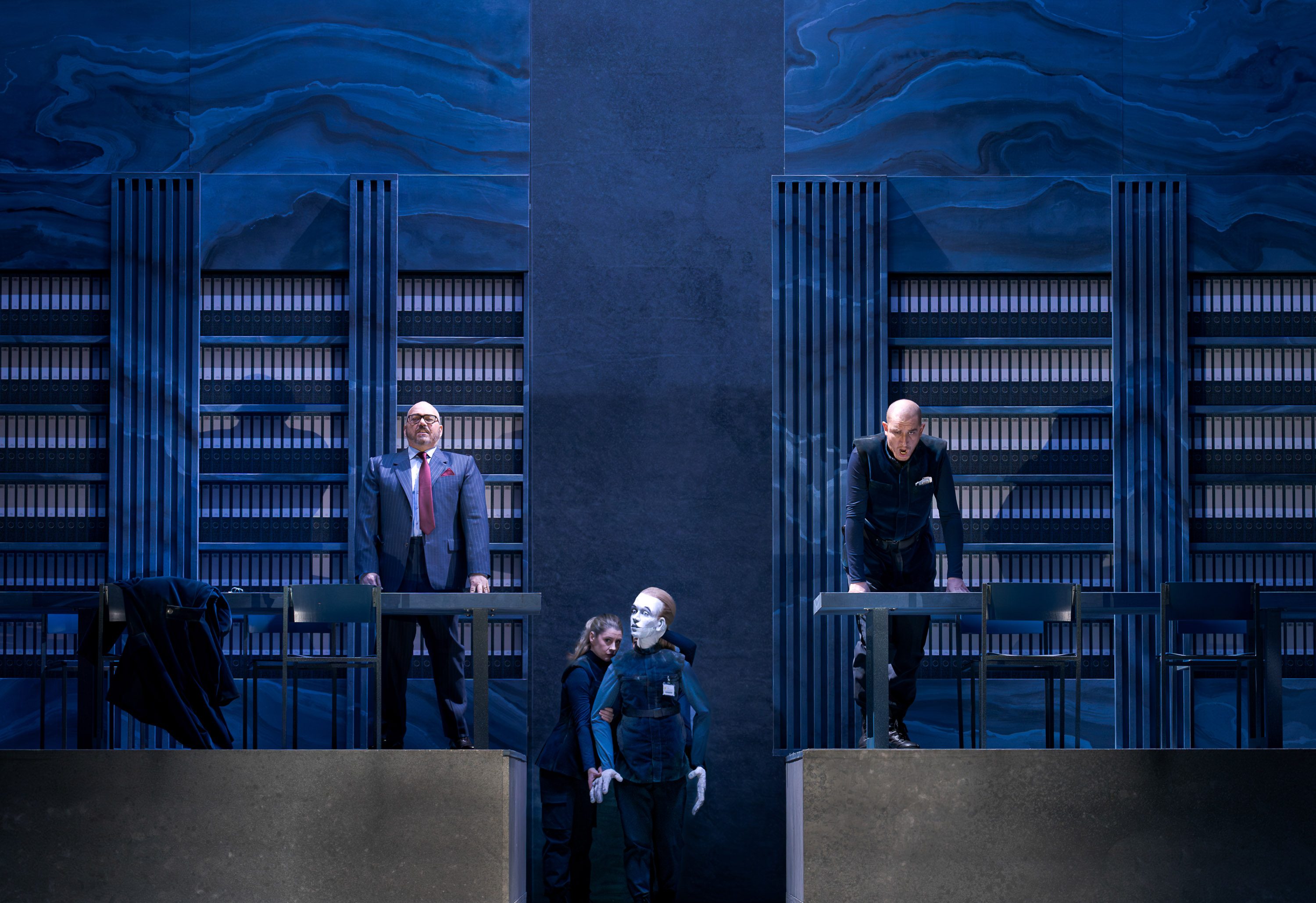 The image depicts a theatrical scene with three characters. Two men stand on elevated platforms, one wearing a suit and the other in a dark outfit, both displaying strong expressions. A woman crouches between them, holding a mannequin-like figure, suggesting a dramatic narrative unfolding in a setting resembling a library.