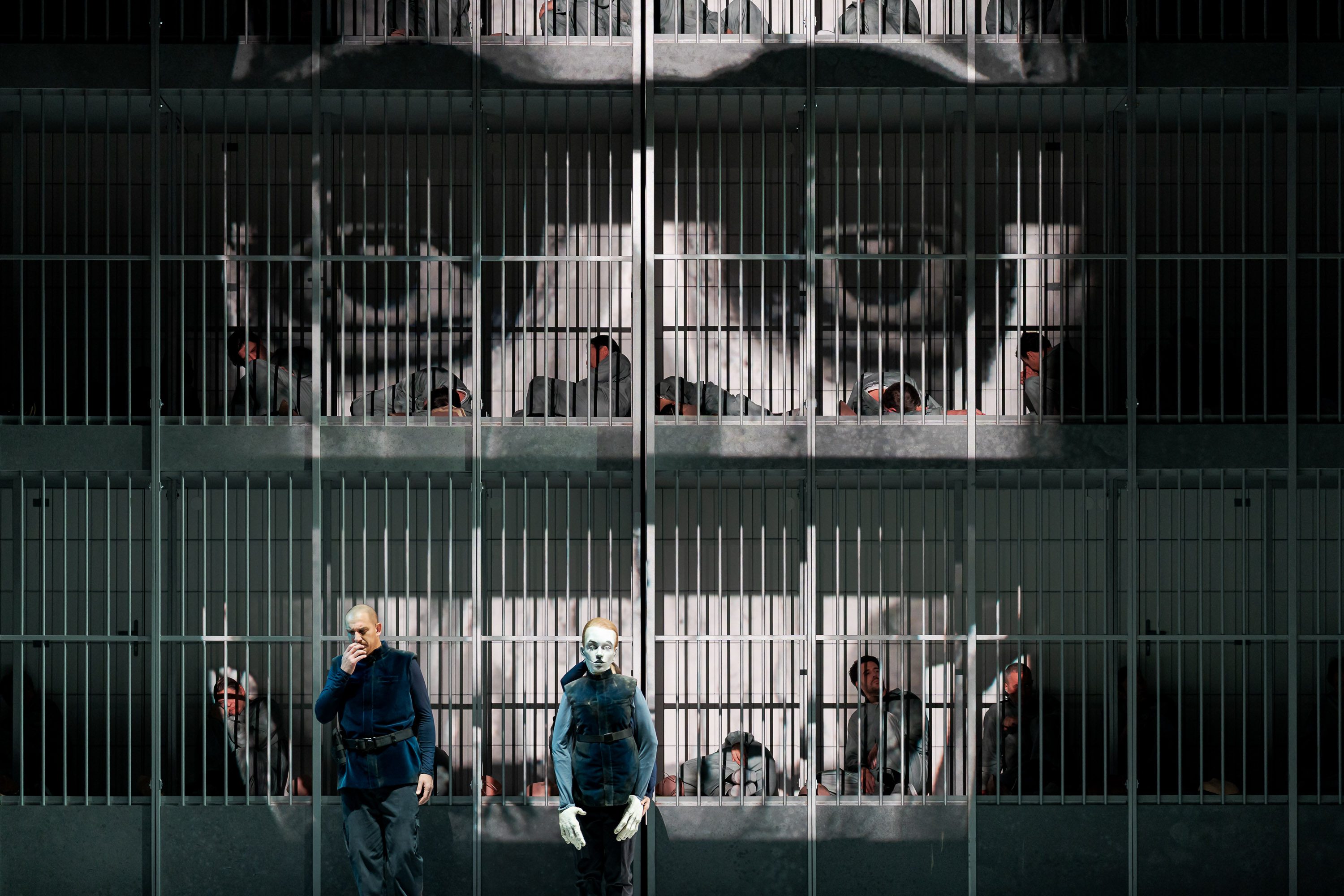 Eine düstere Szene in einer Gefängnisumgebung, in der mehrere Häftlinge hinter Gittern liegen. Ein Mann in blauer Kleidung steht im Vordergrund, neben einer Puppe mit einem ausdruckslosen Gesicht. Die Atmosphäre vermittelt eine starke Gefangenschaft und Isolation.