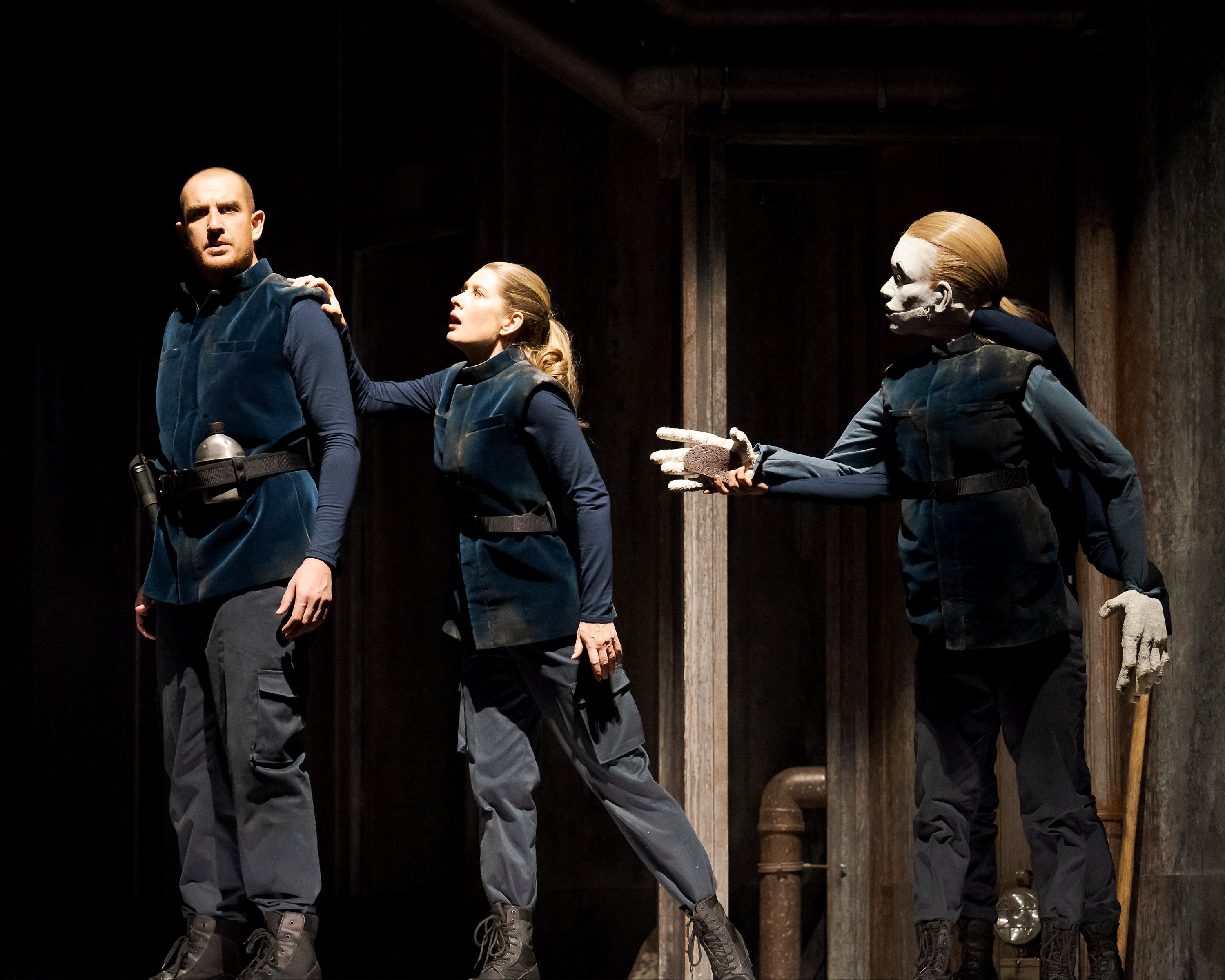 A theatrical scene featuring two actors in dark, modern attire and a puppet. One actor appears concerned, reaching out to the other, while the puppet gestures towards them. The setting is dimly lit, enhancing the tense atmosphere of the moment.