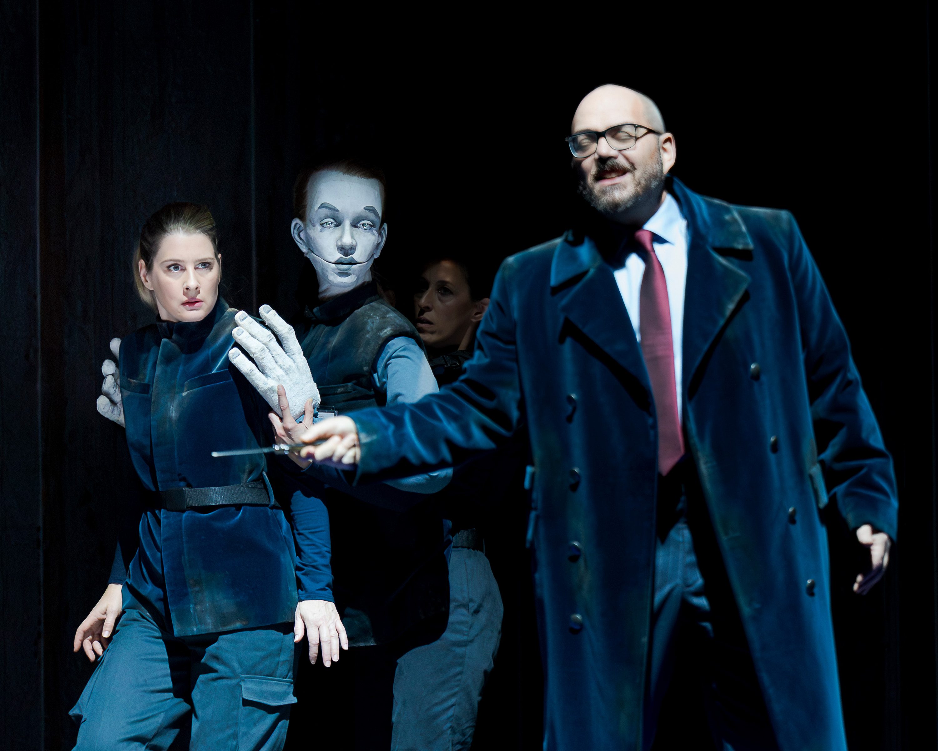 A dramatic scene features a woman in a blue outfit, appearing tense as she stands close to a puppet-like figure with a white face. A man in a dark coat holds a small object and looks intently at the woman, creating a sense of tension and interaction.