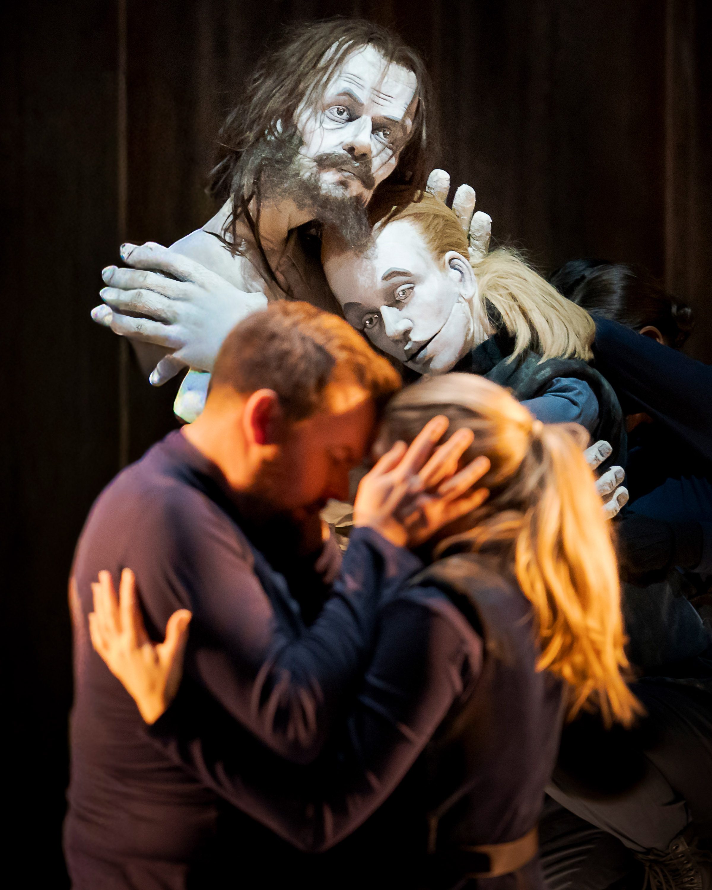 Eine dramatische Szene, in der ein lebensgroßes, skulpturales Gesicht zwischen zwei Menschen steht, die sich innig umarmen. Die Emotionen sind stark, mit einem Fokus auf Verbindung und Nähe. Der Hintergrund ist dunkel, was die Figuren im Vordergrund hervorhebt.