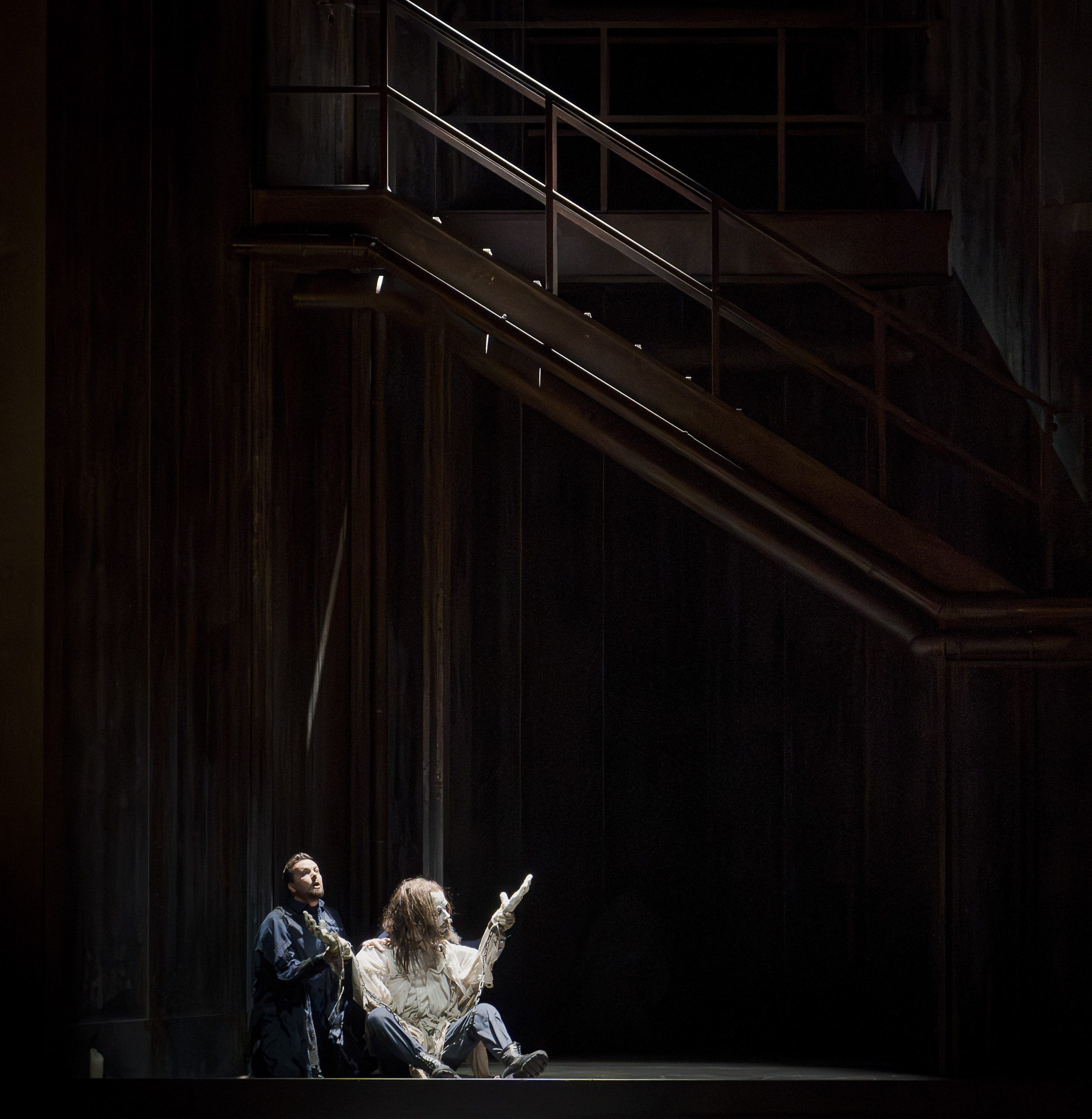 A dimly lit stage shows two figures seated on the floor; one is looking up and pointing, while the other gazes intently. The background features a large staircase, enhancing the dramatic atmosphere of the scene.
