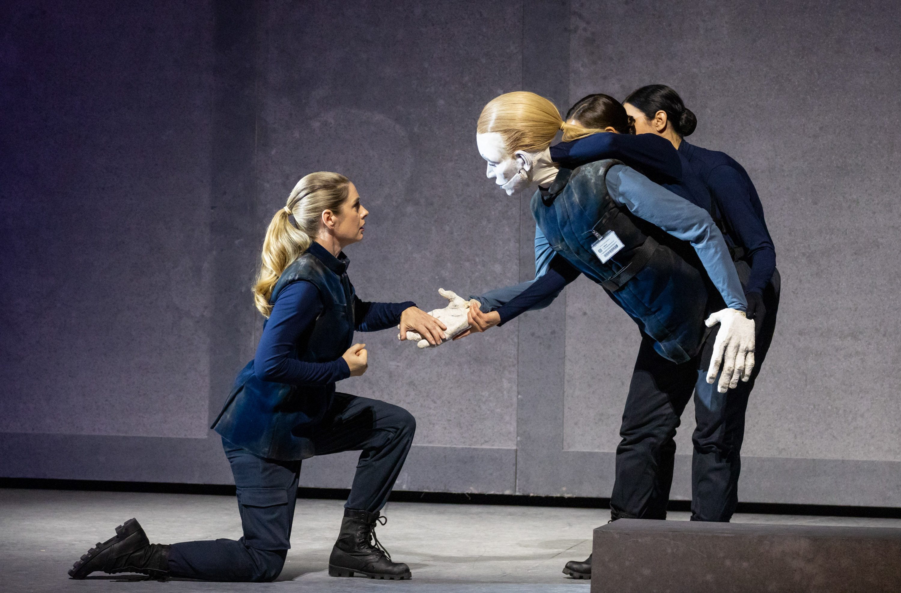 A performer kneels, reaching out to shake hands with a humanoid figure that has a painted face and is being supported by another performer. The scene conveys a moment of connection or agreement between them, set against a simple gray backdrop.