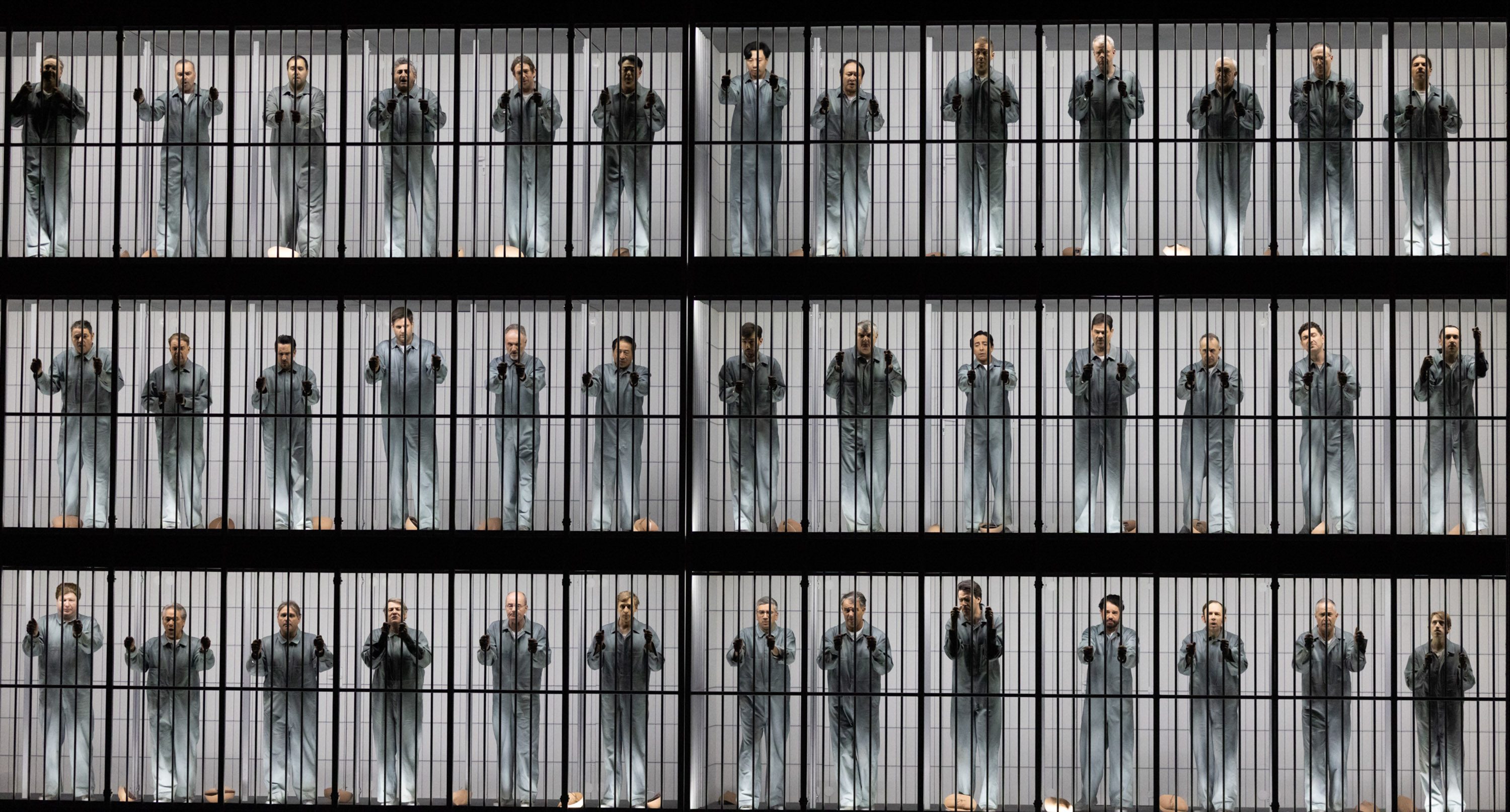 The image features multiple individuals dressed in gray prison uniforms, standing behind bars on two tiers. Each person poses with their hands on the bars, conveying a sense of confinement and shared experience. The background is minimal, highlighting their expressions and body language.