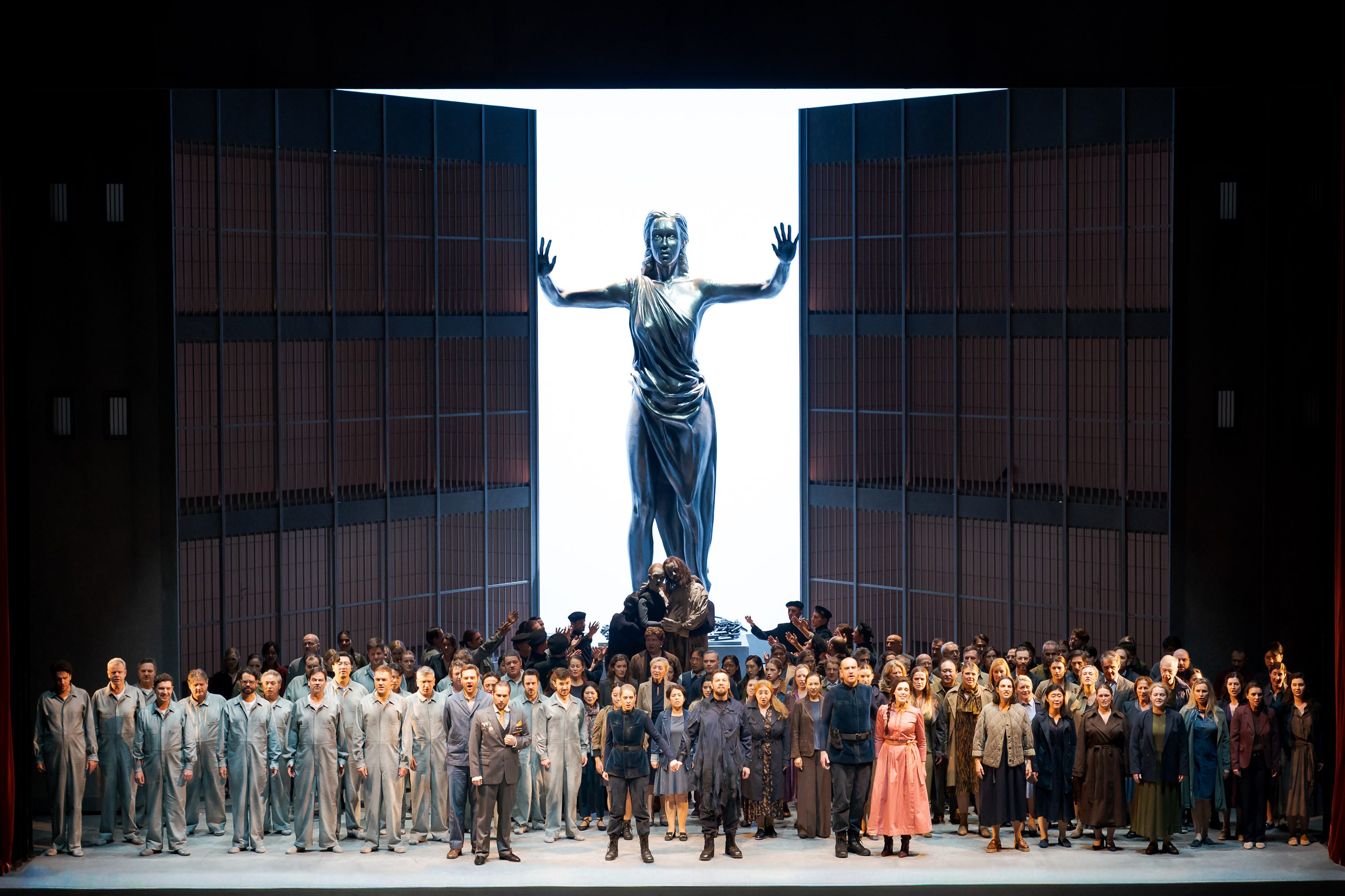 A large group of performers stands on stage, dressed in various costumes, in front of a towering statue of a woman with outstretched arms. The background features large, open doors revealing a bright light, symbolizing hope or freedom. The atmosphere suggests a dramatic performance.