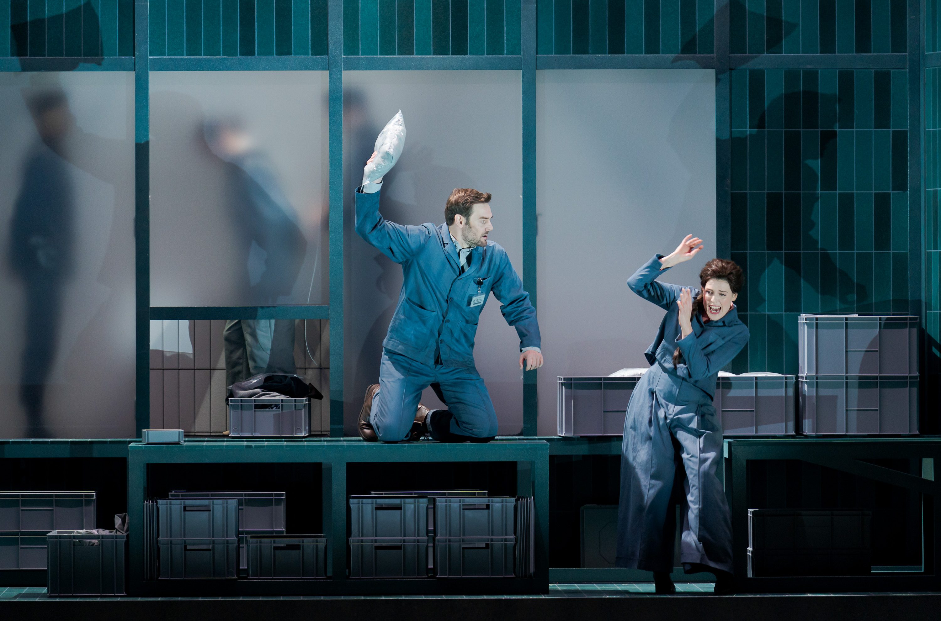 A man in a blue uniform kneels on a platform, raising a pillow as if to throw it. A woman, also in blue, recoils in surprise, shielding her face. They are in a set with storage boxes, creating a playful or dramatic scene.