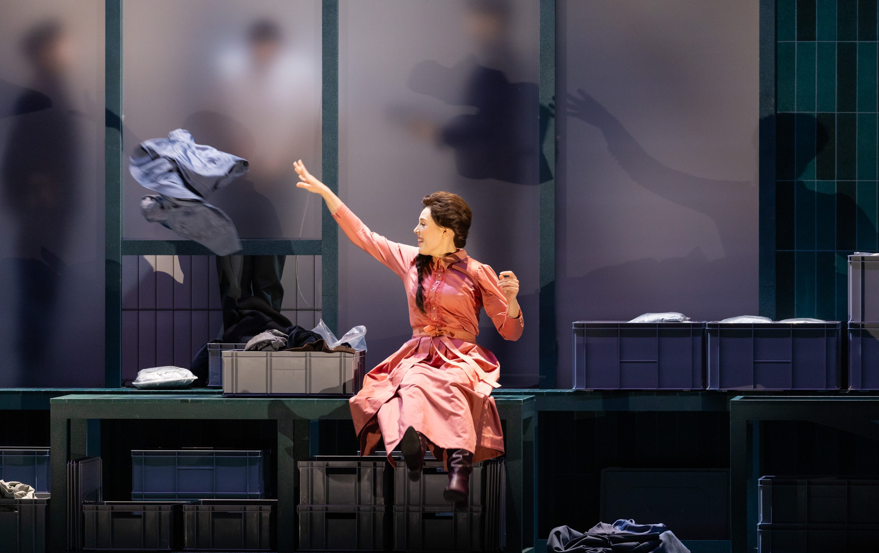 A woman in a pink dress sits on a table, reaching out as she throws a piece of clothing. Behind her, shadowy figures can be seen through frosted panels. The scene captures an energetic moment in a theatrical setting, emphasizing the woman's expressive actions.