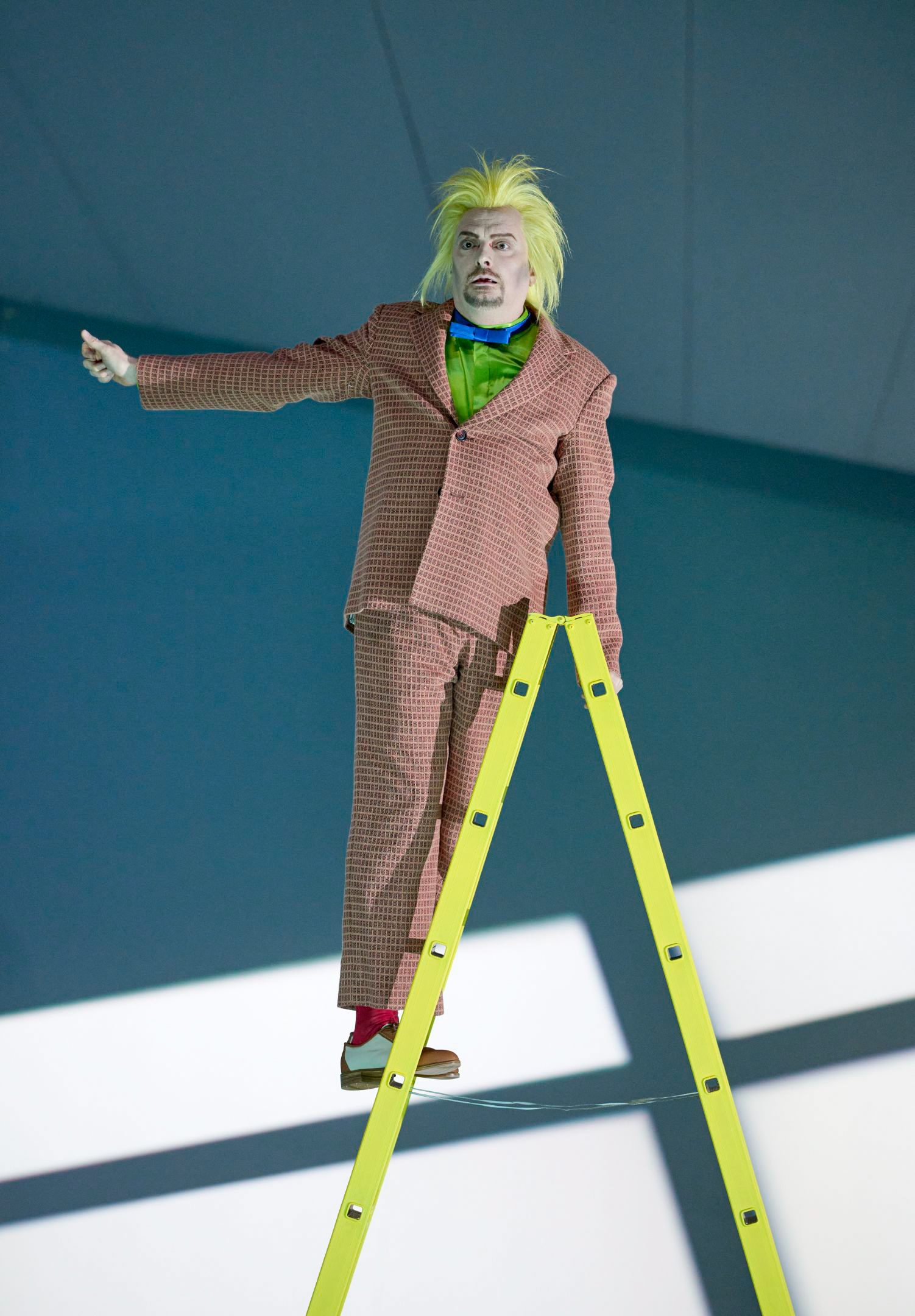 Eine Figur mit gelber Perücke und orangefarbenem Anzug steht auf einer Leiter und zeigt mit ausgestrecktem Arm in die Ferne.