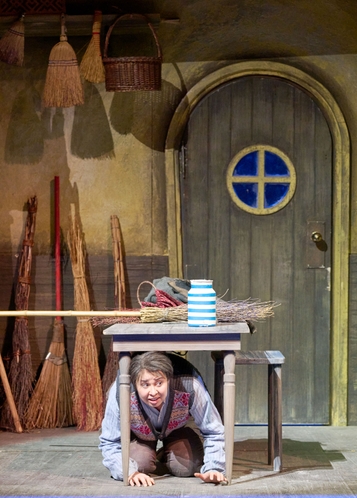 Eine Person hockt mit ängstlichem Blick unter einem Tisch auf der Bühne in einer rustikal gestalteten Stube mit mehreren Besen im Hintergrund.