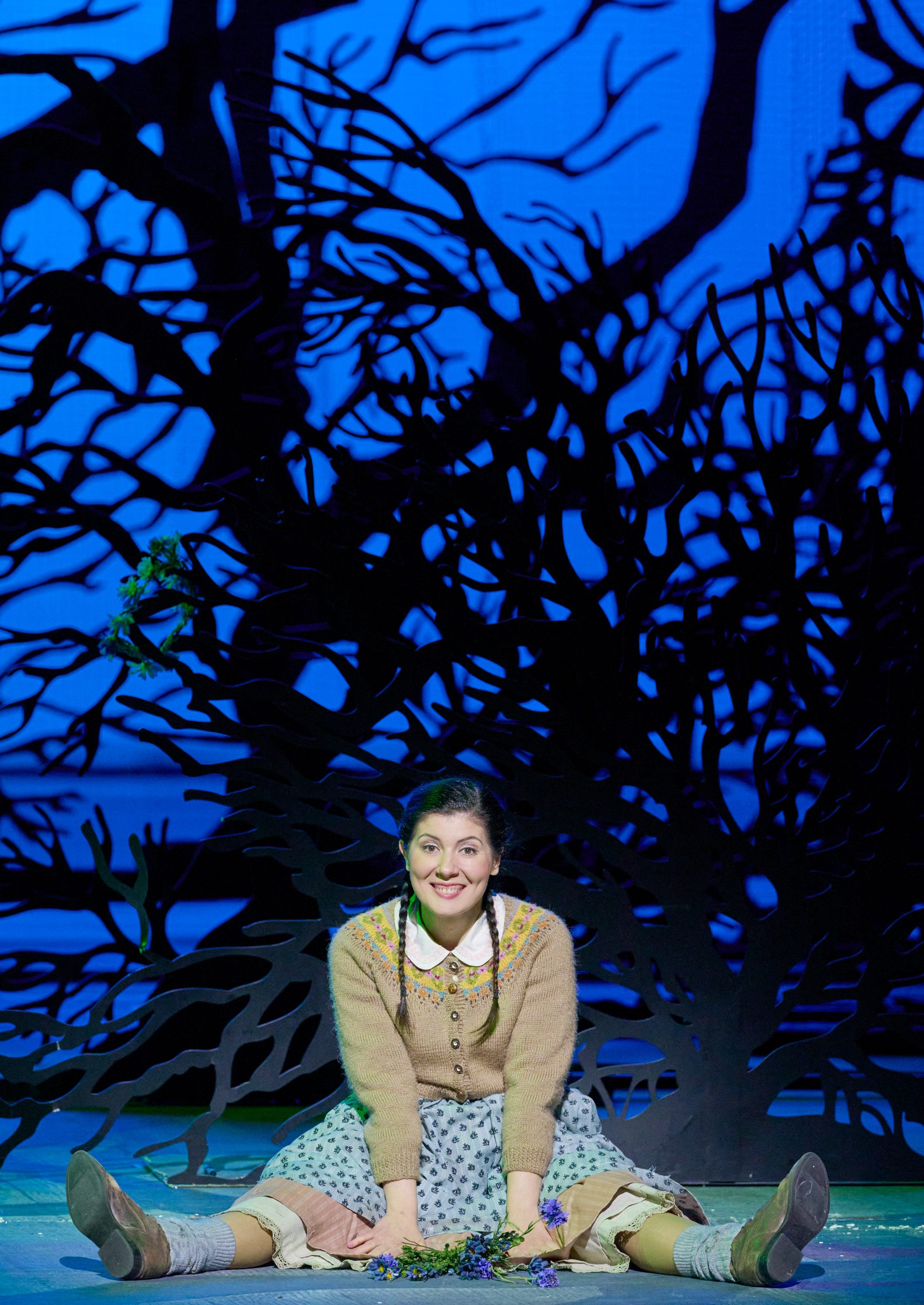 Ein junges Mädchen in einem altmodischen Kleid sitzt fröhlich mit gespreizten Beinen auf der Bühne vor einer märchenhaften Kulisse mit dunklen Baumumrissen.