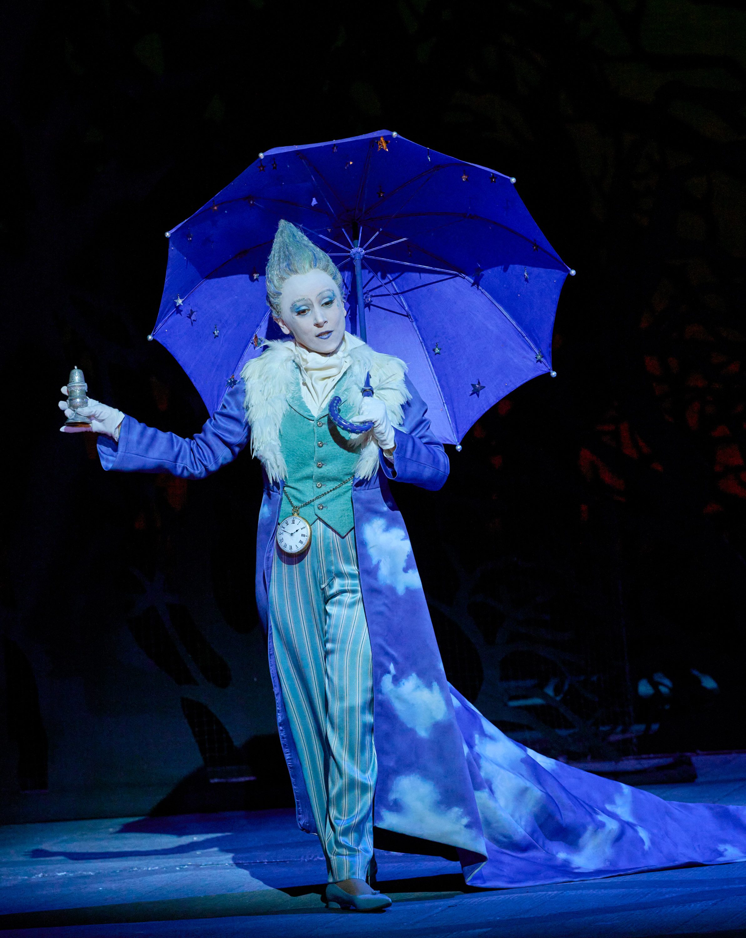 Eine märchenhaft gekleidete Figur mit hellblauer Haut und aufgespanntem Schirm steht auf der Bühne und hält eine kleine Glaskugel in der Hand.