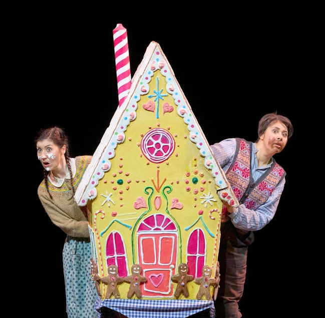 Zwei Kinder blicken neugierig hinter einem bunten, märchenhaften Lebkuchenhaus hervor, das auf der Bühne steht.