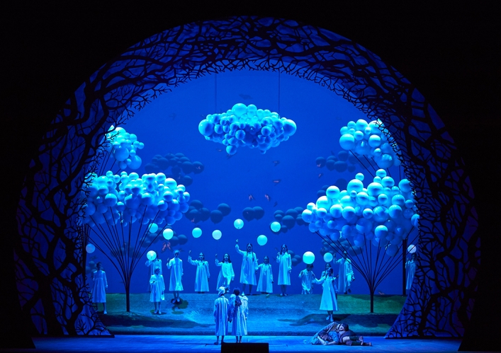 Eine märchenhafte Bühnenlandschaft in Blau mit leuchtenden Bäumen, schwebenden Wolken und Figuren in weißen Gewändern, die feierlich versammelt sind.