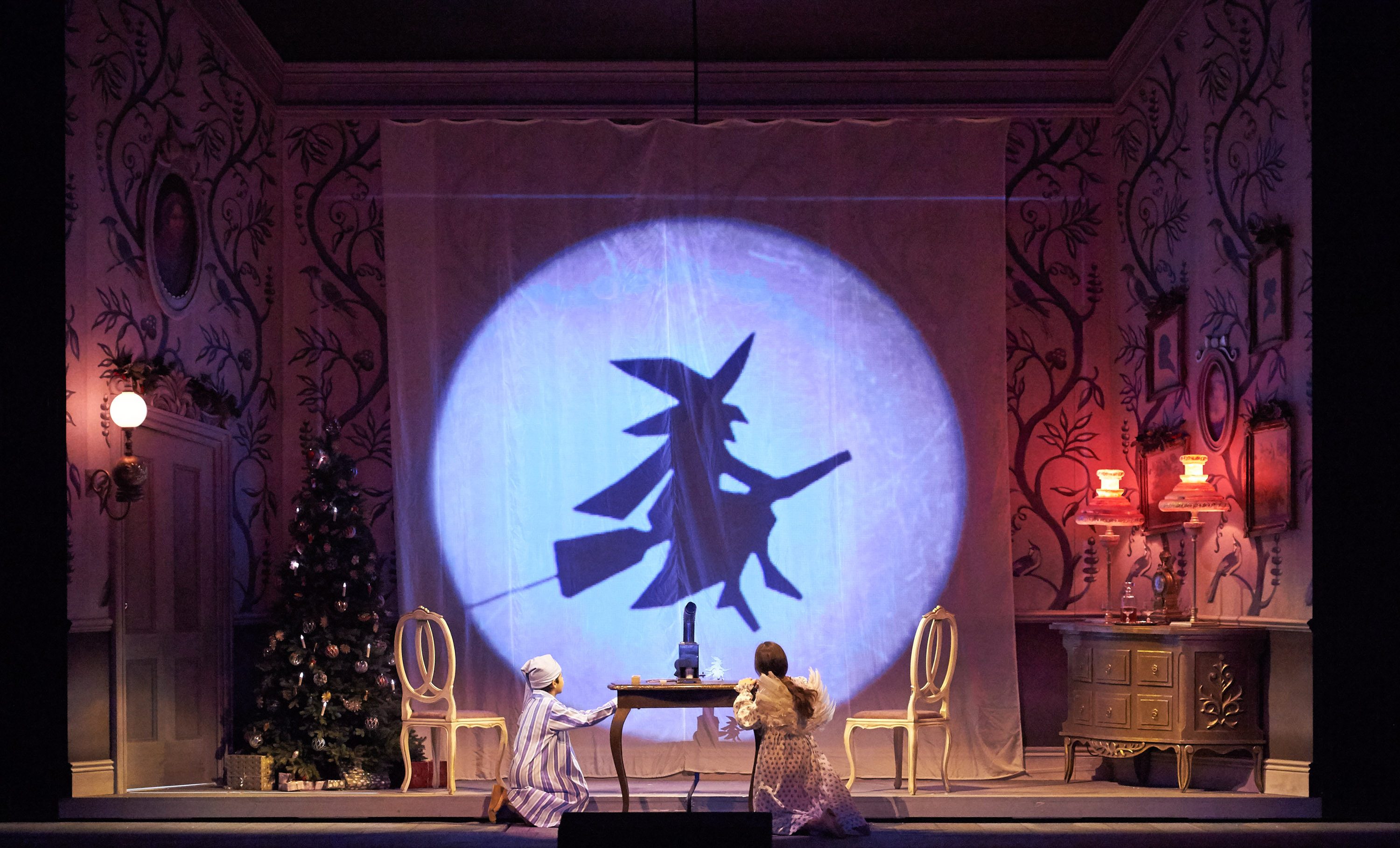 Zwei Kinder sitzen an einem Tisch in einem festlich dekorierten Raum, während auf einem Vorhang die Schattenprojektion einer fliegenden Hexe zu sehen ist.