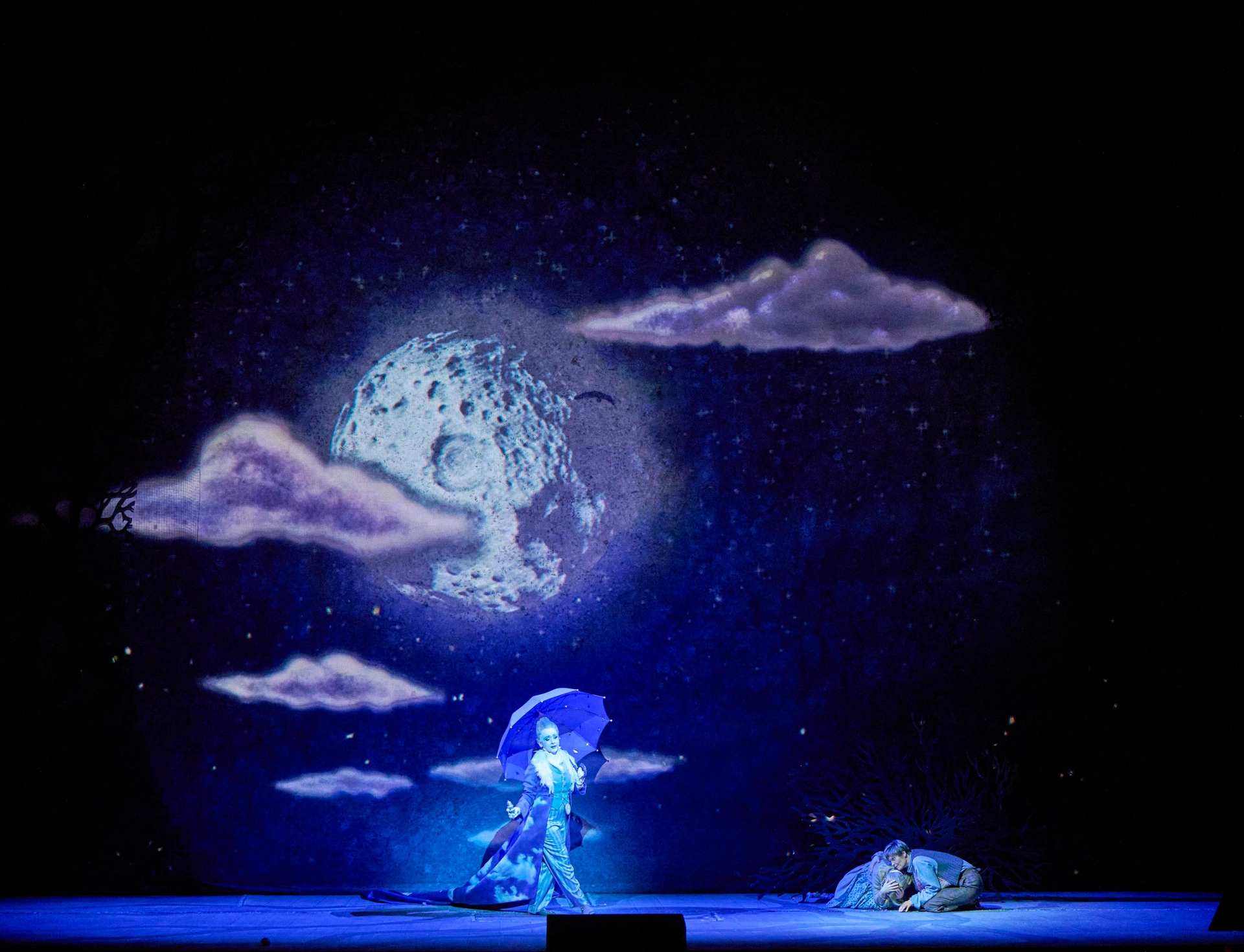 A performer in a flowing blue outfit holds a purple umbrella against a backdrop of a moon and clouds. Another figure is curled up on the ground. The scene is illuminated with soft blue and purple lighting, creating a dreamlike atmosphere.