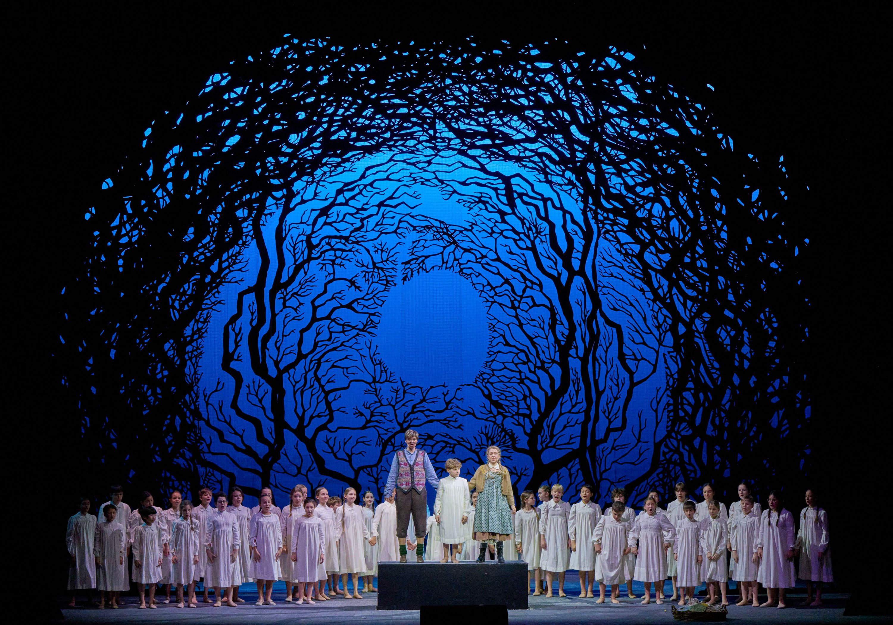 A group of children and two main characters stand on stage, surrounded by a dramatic blue backdrop featuring intricate black tree branches. They are dressed in white nightgowns, conveying a sense of innocence and wonder as they perform together.
