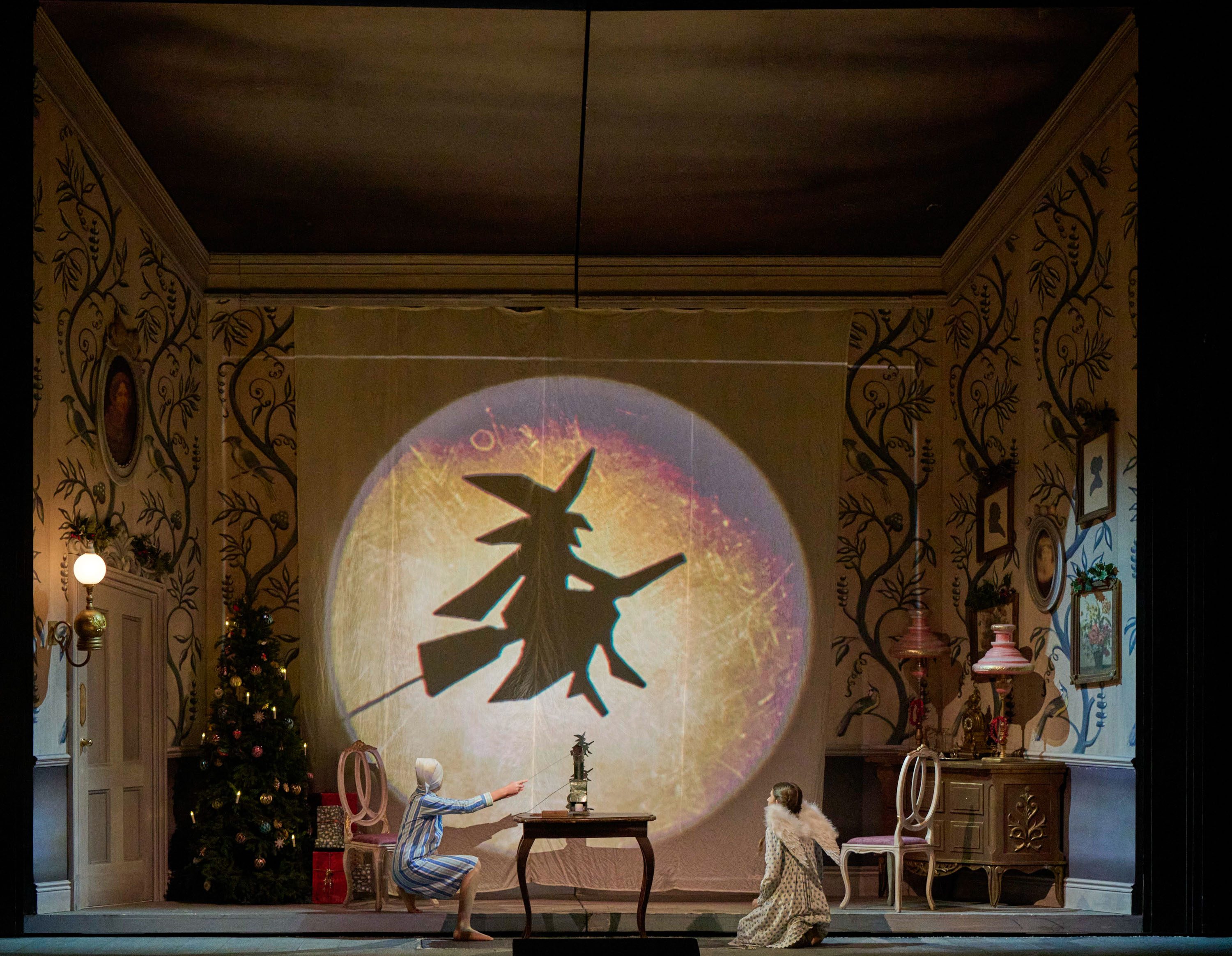 The image depicts two children in a warmly decorated room, captivated by a large shadow projection of a witch on a backdrop. One child reaches towards the shadow, while the other sits on the floor in a whimsical outfit, emphasizing a sense of wonder and enchantment.