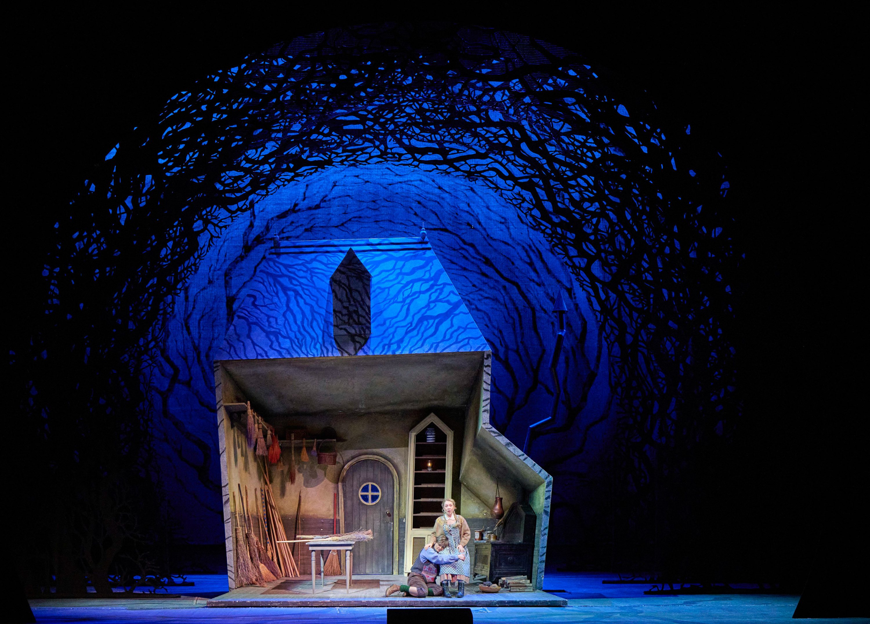 A stage scene features a tilted house with a door and window, surrounded by dark, leafy silhouettes. Two figures are seated in front of the house, one kneeling and the other seated on a small stool, creating a dramatic atmosphere illuminated by blue light.