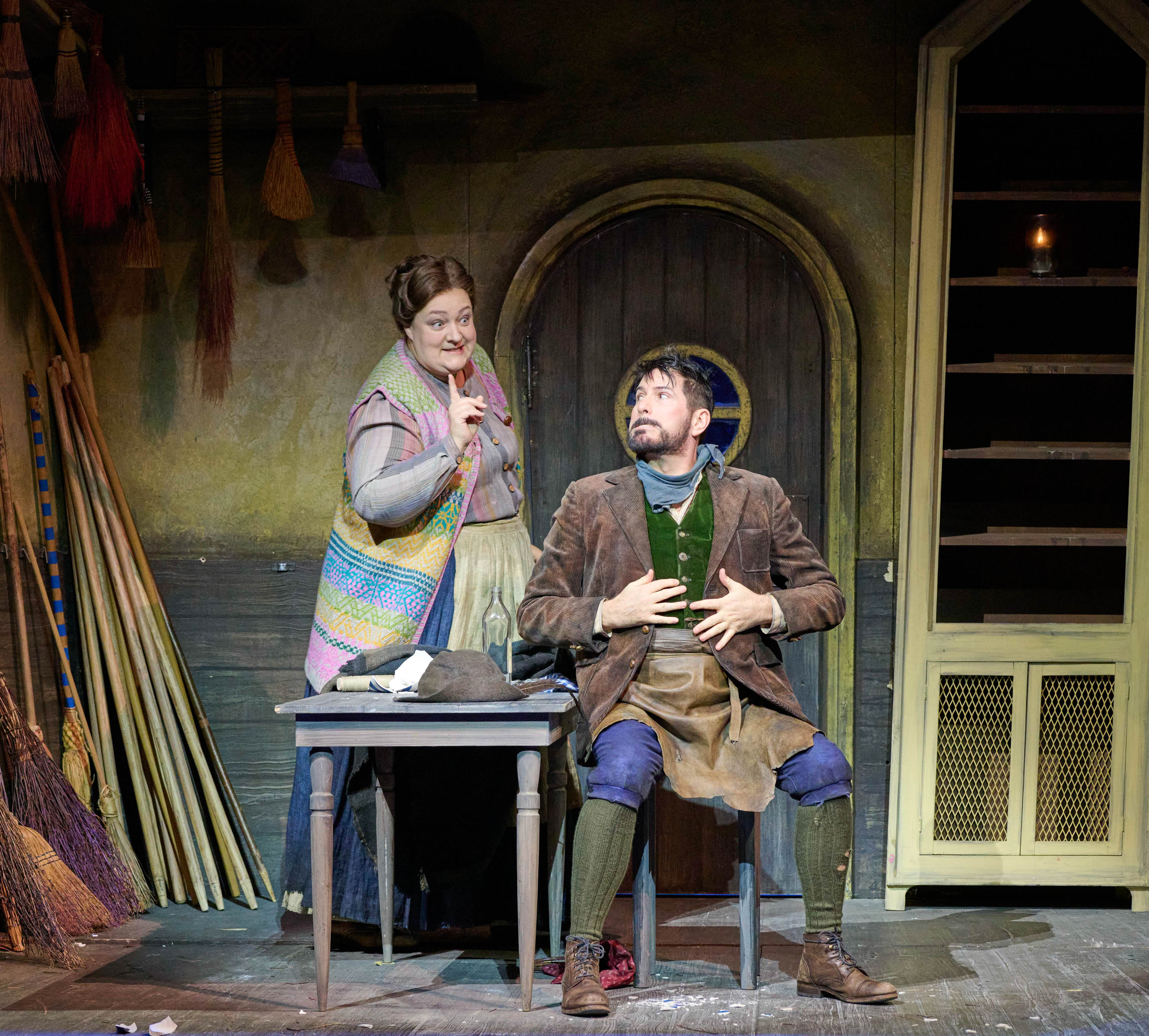 A woman gestures playfully while standing beside a seated man who looks surprised. They are in a rustic setting with brooms hanging on the wall. The woman wears a colorful vest over a long skirt, while the man is dressed in a brown jacket and apron.