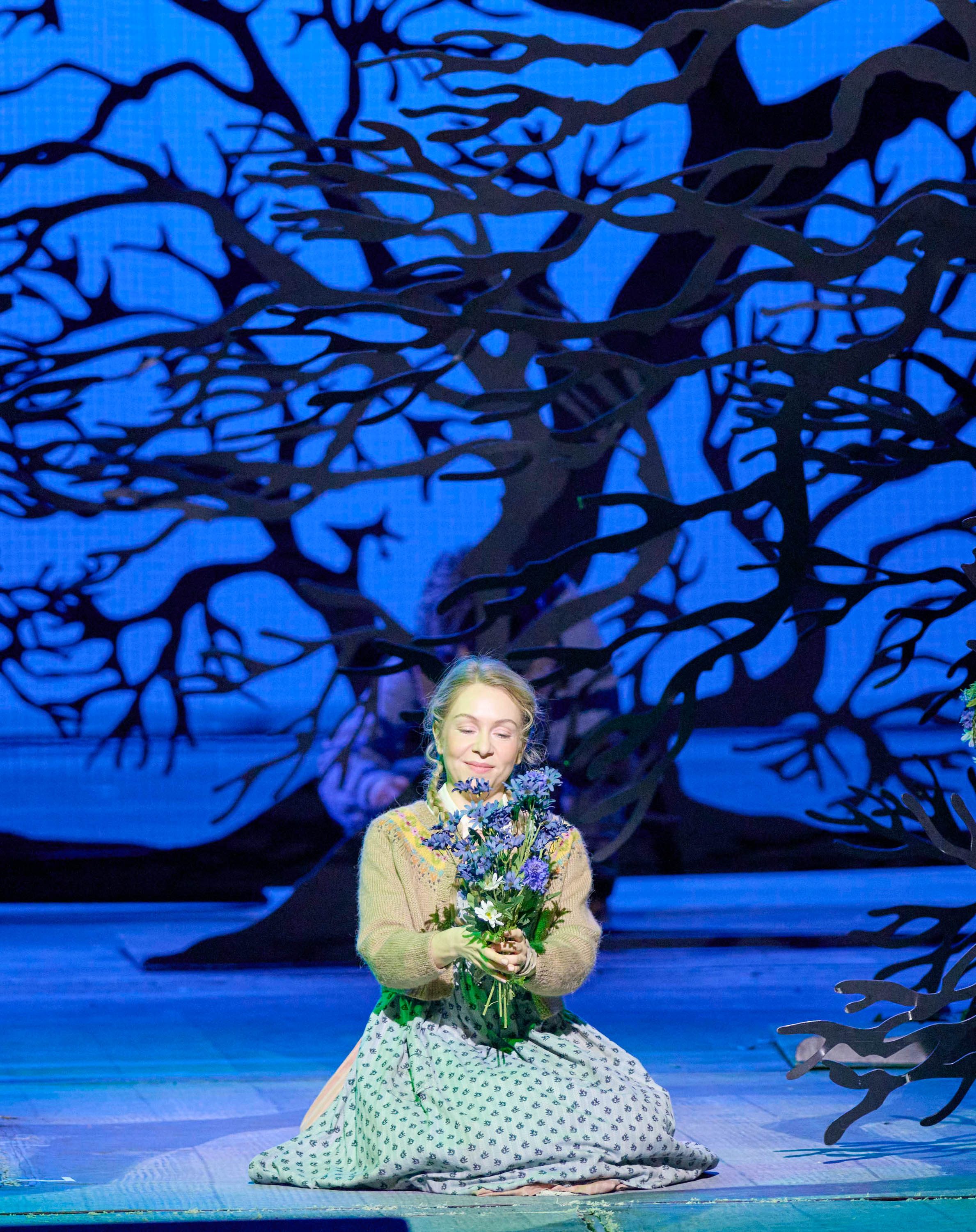 A young woman kneels on stage, holding a bouquet of flowers close to her heart. She has a serene expression, surrounded by dark, intricately cut tree silhouettes against a bright blue background, creating a whimsical, theatrical atmosphere.