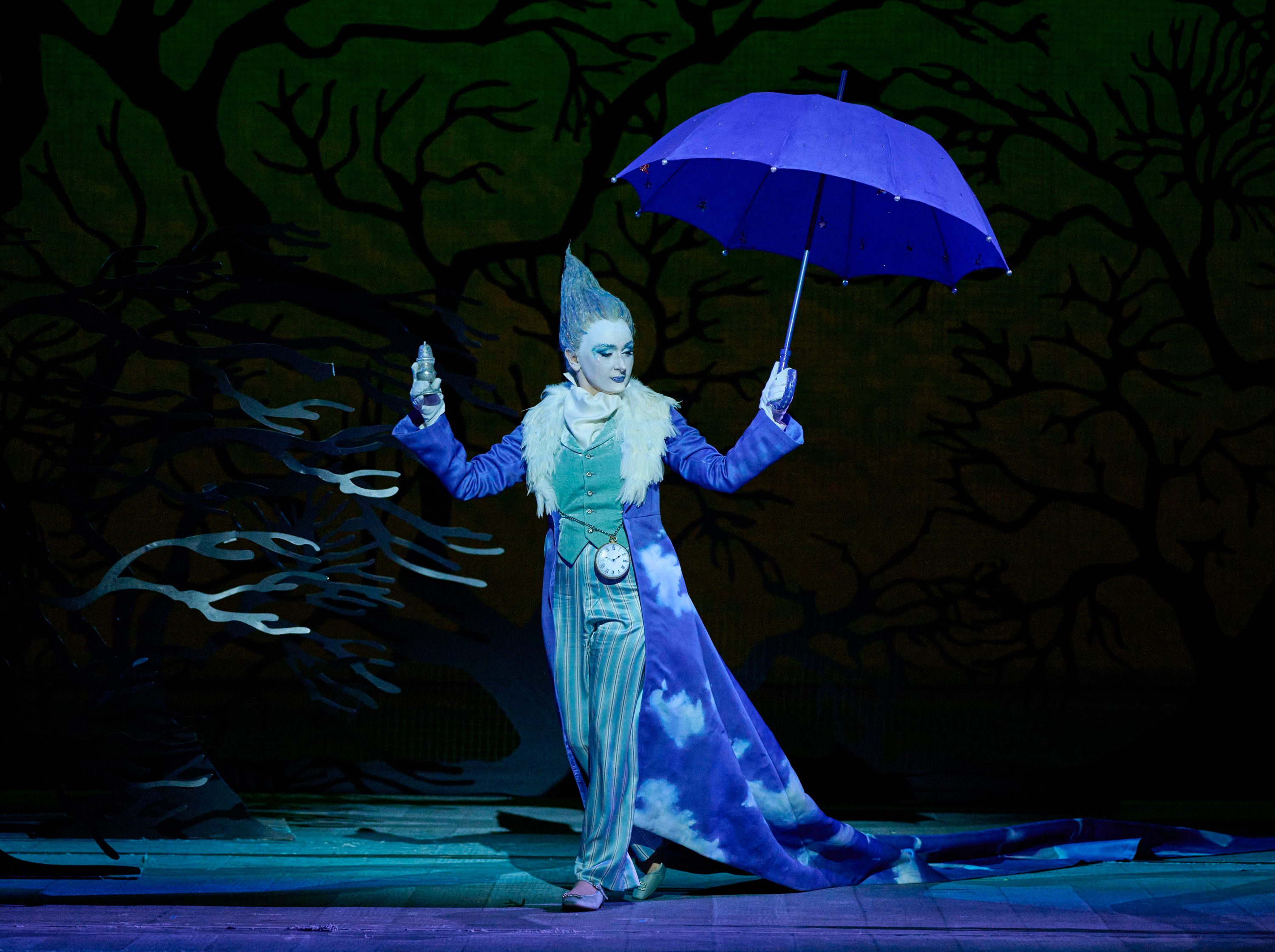 A performer dressed in a blue and white costume, resembling a whimsical character, holds a purple umbrella in one hand and a shimmering bottle in the other. The background features dark, bare trees, suggesting a mystical atmosphere.