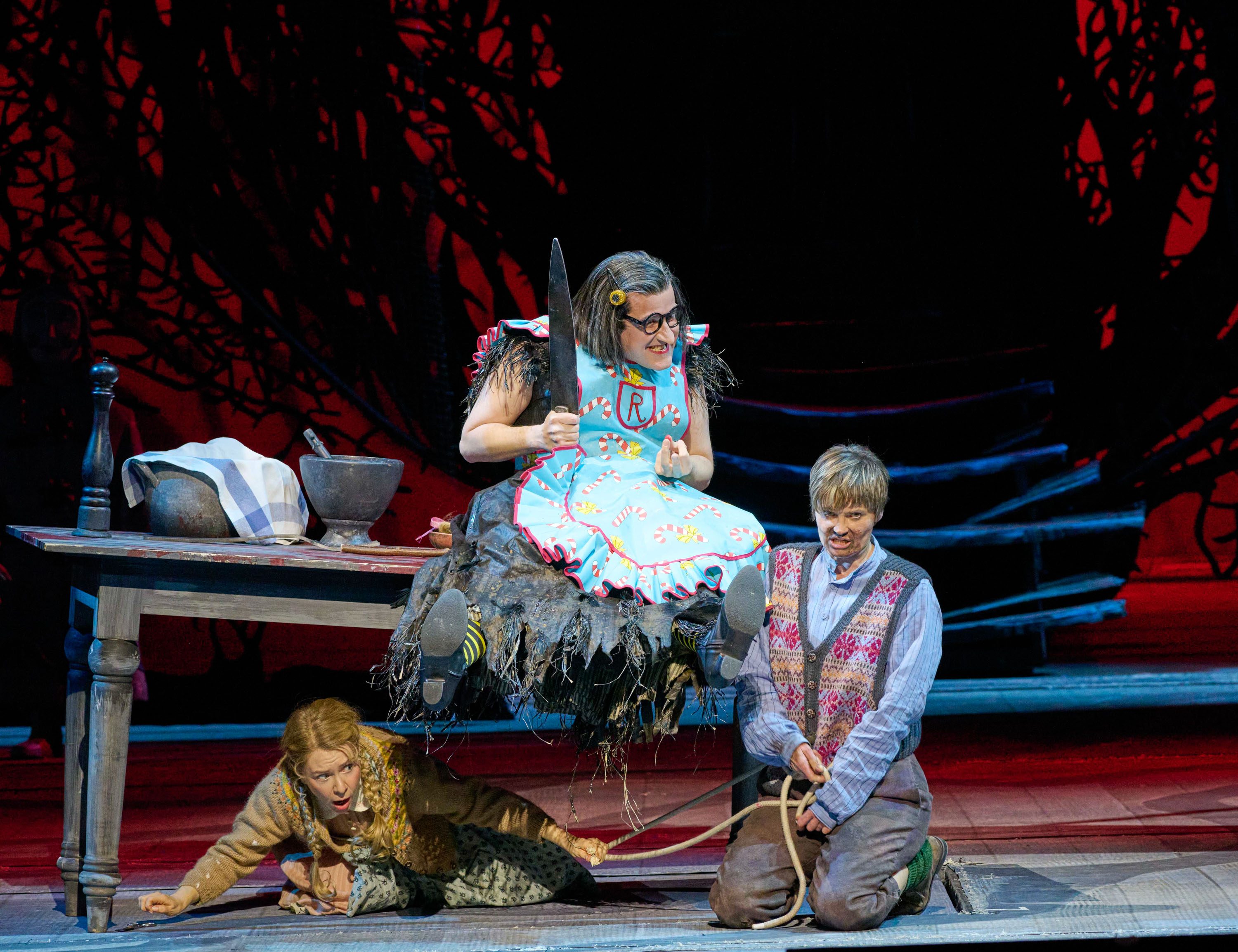 A character in a whimsical blue dress with candy patterns sits on a table holding a prop knife, while two other characters are on the ground: one appears frightened and hiding, and the other is kneeling with a rope. The background features dark, stylized trees.