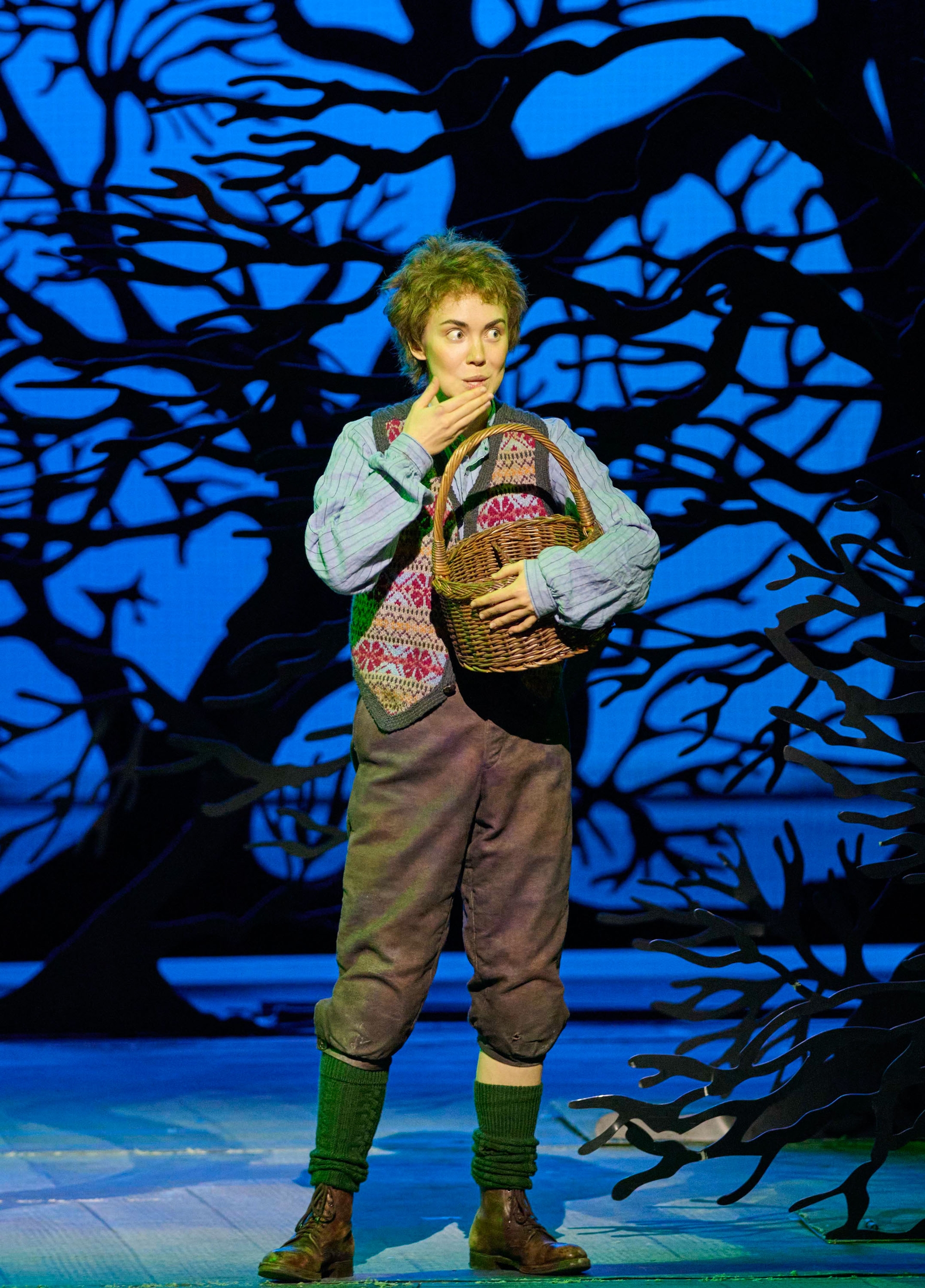 A young performer stands in a theatrical setting, holding a wicker basket. They have a surprised expression, wearing a colorful, patterned vest over a long-sleeved shirt and loose trousers. Dark, silhouetted tree branches frame the background, creating an enchanted forest atmosphere.
