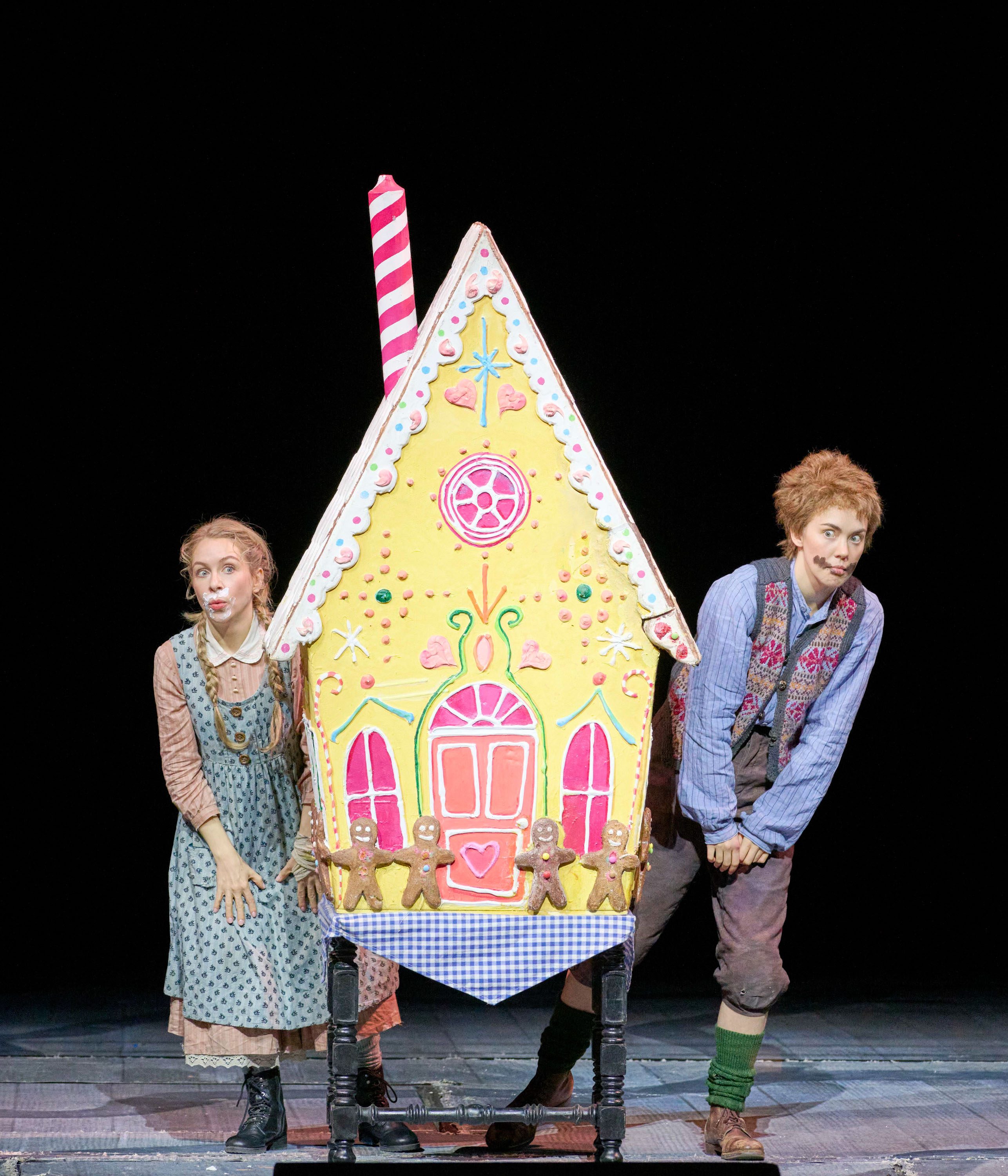 Two actors stand beside a colorful, oversized gingerbread house decorated with candy and icing. One actor, a girl, has a surprised expression, while the boy next to her is playfully peeking out. The backdrop is dark, highlighting their whimsical costumes and the vibrant house.