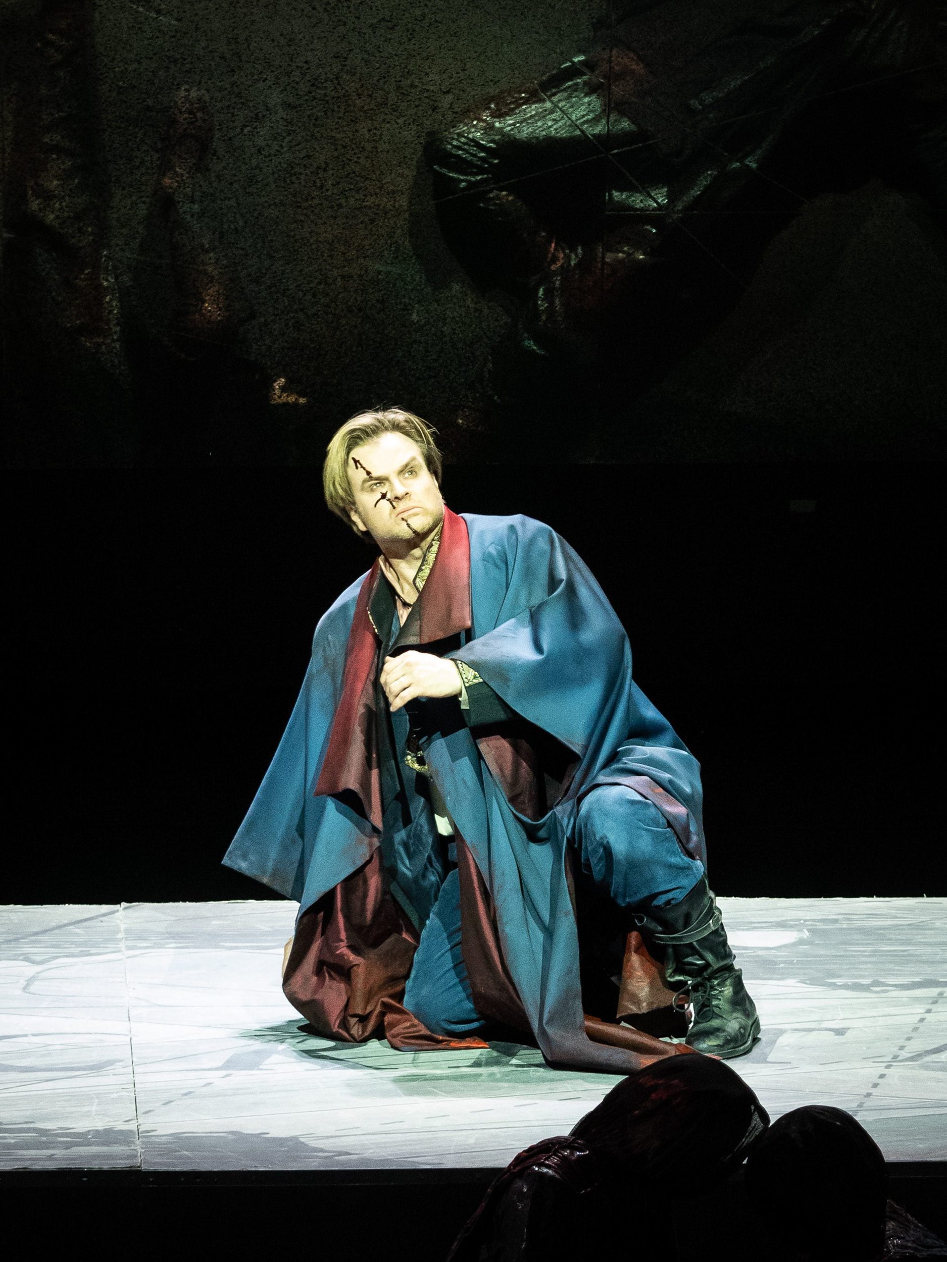 Ein Mann in blauem Gewand bückt sich vor einem dunklen Hintergrund; er blickt entschlossen nach vorne.