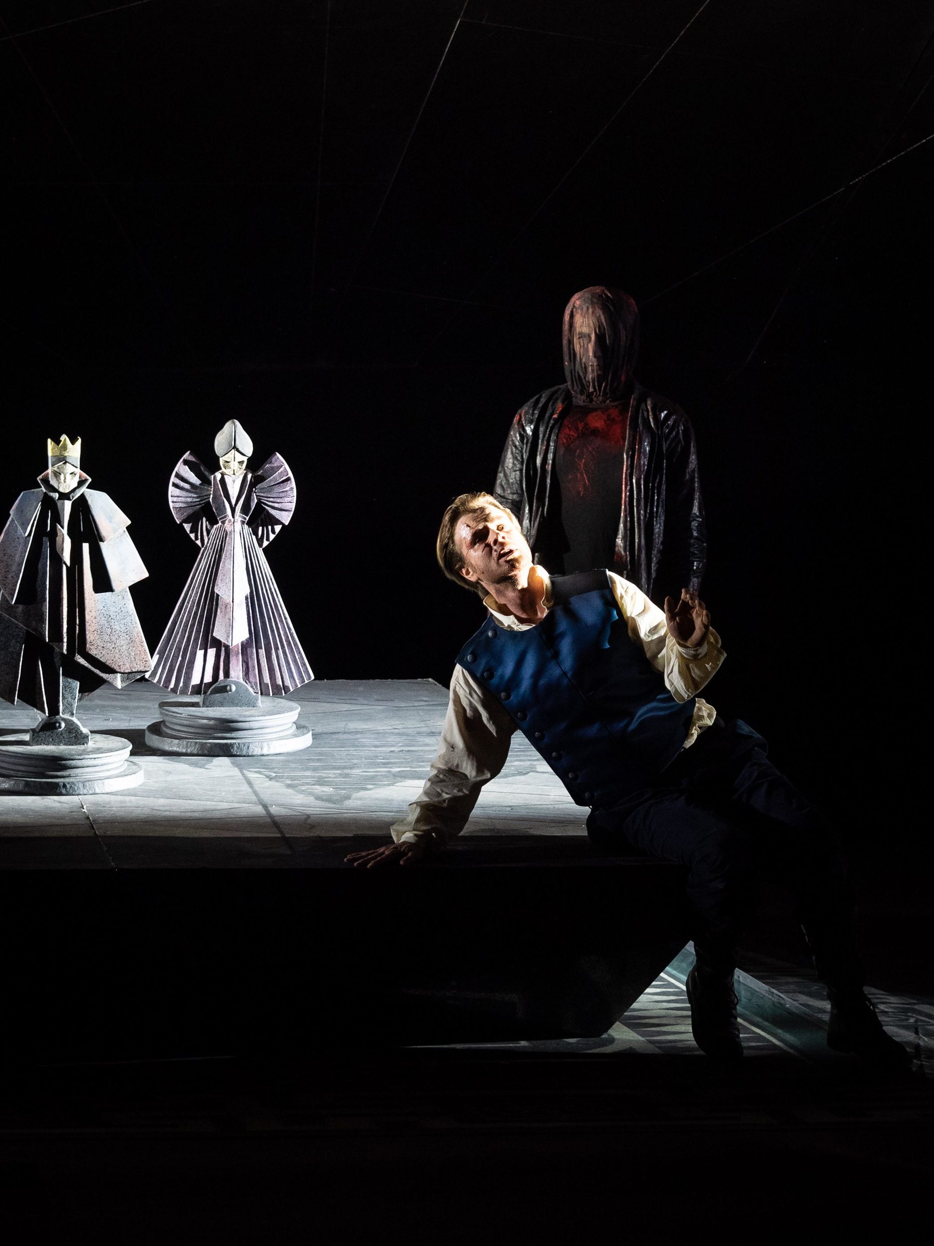 Ein Mann liegt auf einer dunklen Bühne; im Hintergrund sind zwei helle Statuen, sowie eine weitere Person, die ihn bedrohlich ansieht.