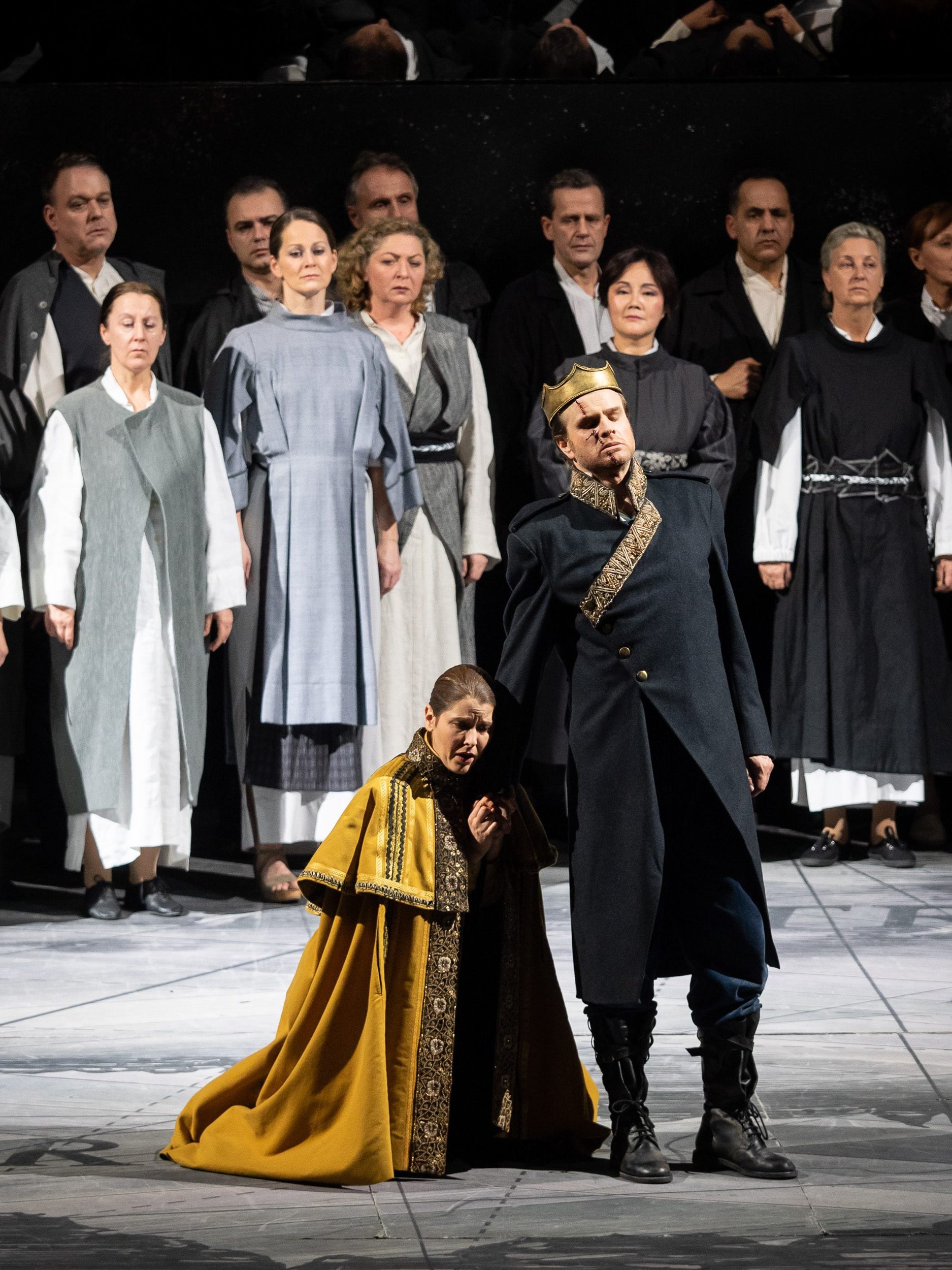 Ein Mann in dunklem Gewand und goldener Krone steht neben einer am Boden knienden Frau, die sich an ihm festhält. Im Hintergrund stehen mehrere Personen in schlichter Kleidung.