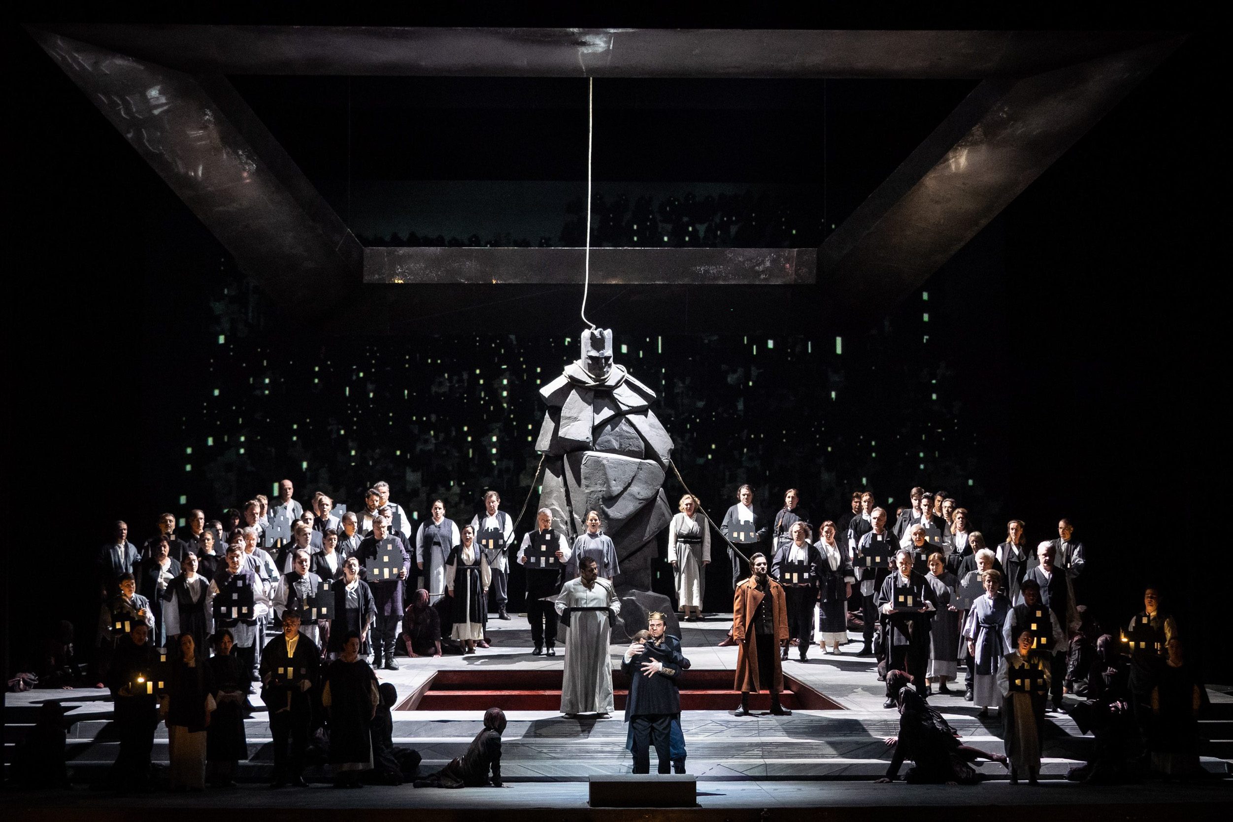 Personen stehen in Kostümen auf der Bühne, die in düsteren Farben gehalten ist. Im Zentrum steht eine große, abstrakte, skulpturale Figur