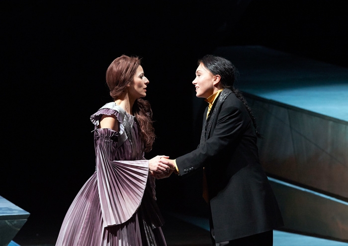 Zwei Personen stehen sich auf der Bühne gegenüber und halten sich an den Händen – die eine in einem historischen Kleid, die andere in modernerem Anzug.