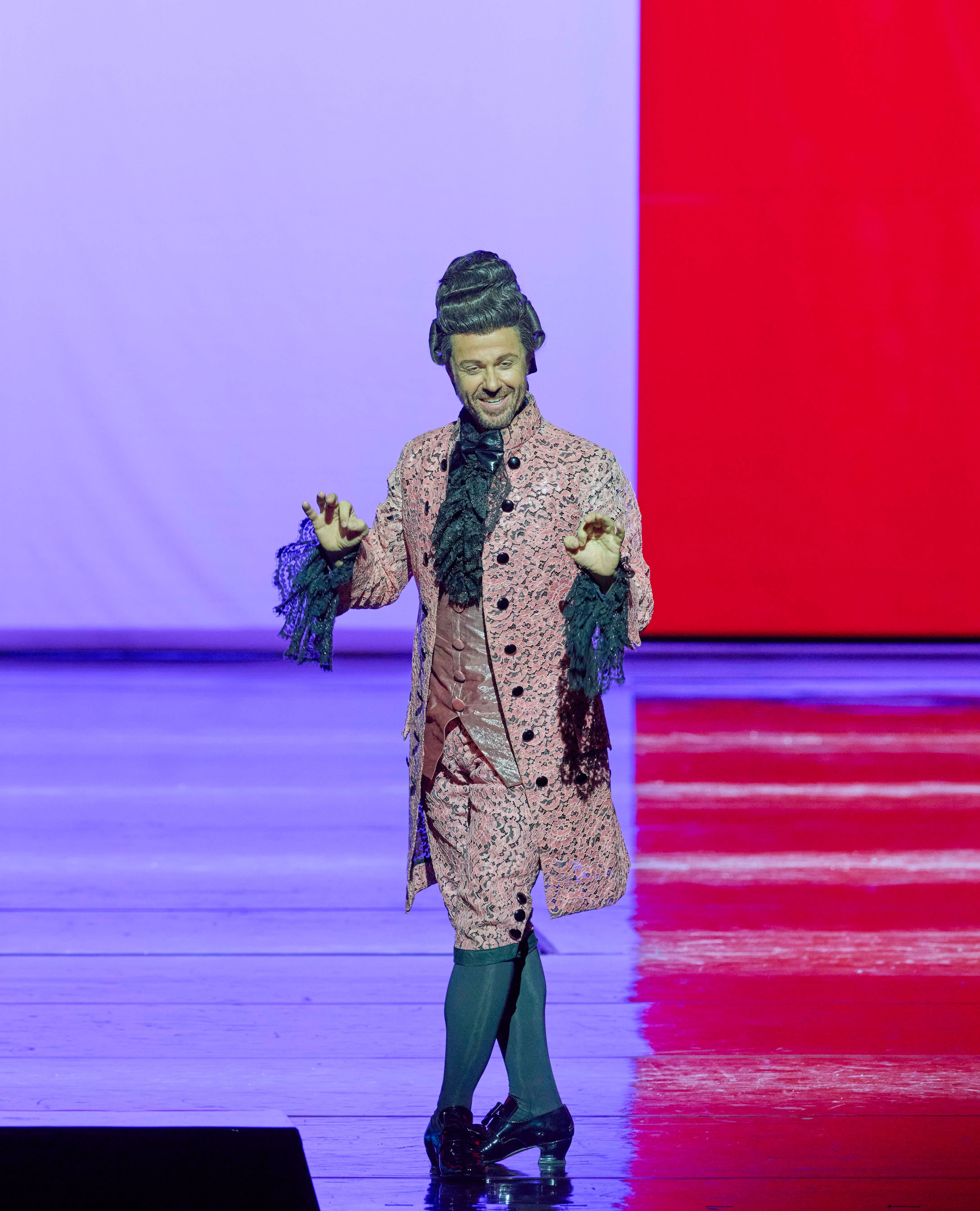 A performer dressed in an elaborate pink costume with lace and ruffles, featuring a large hairstyle, stands on a brightly lit stage. They appear joyful and are gesturing with their hands, engaging the audience. The backdrop has vibrant abstract colors.