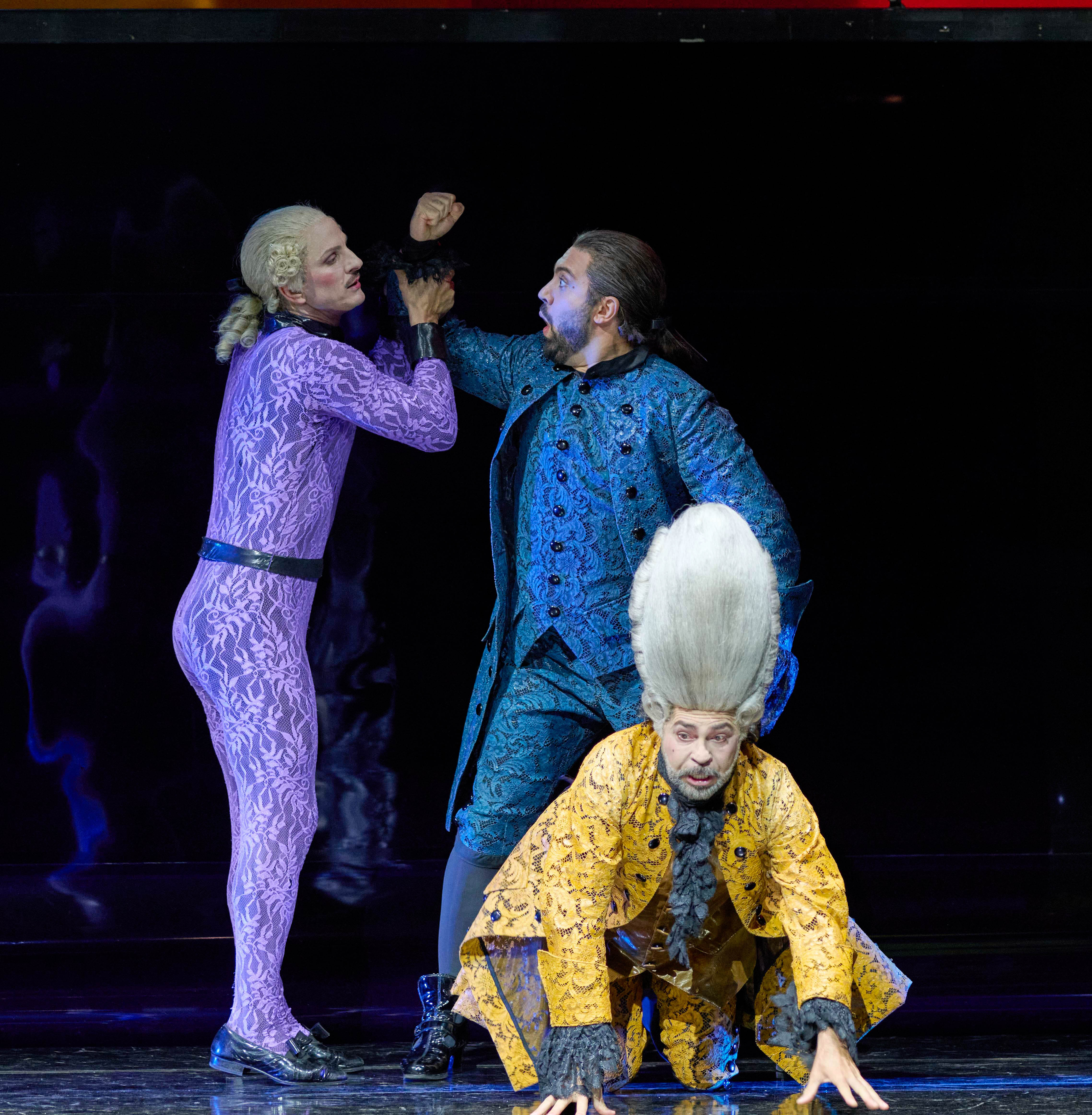 Three performers are on stage in a dramatic scene. One character in a purple outfit confronts another in a blue coat, while a third character in a yellow costume crouches nearby, observing the interaction. The atmosphere conveys tension and intrigue.
