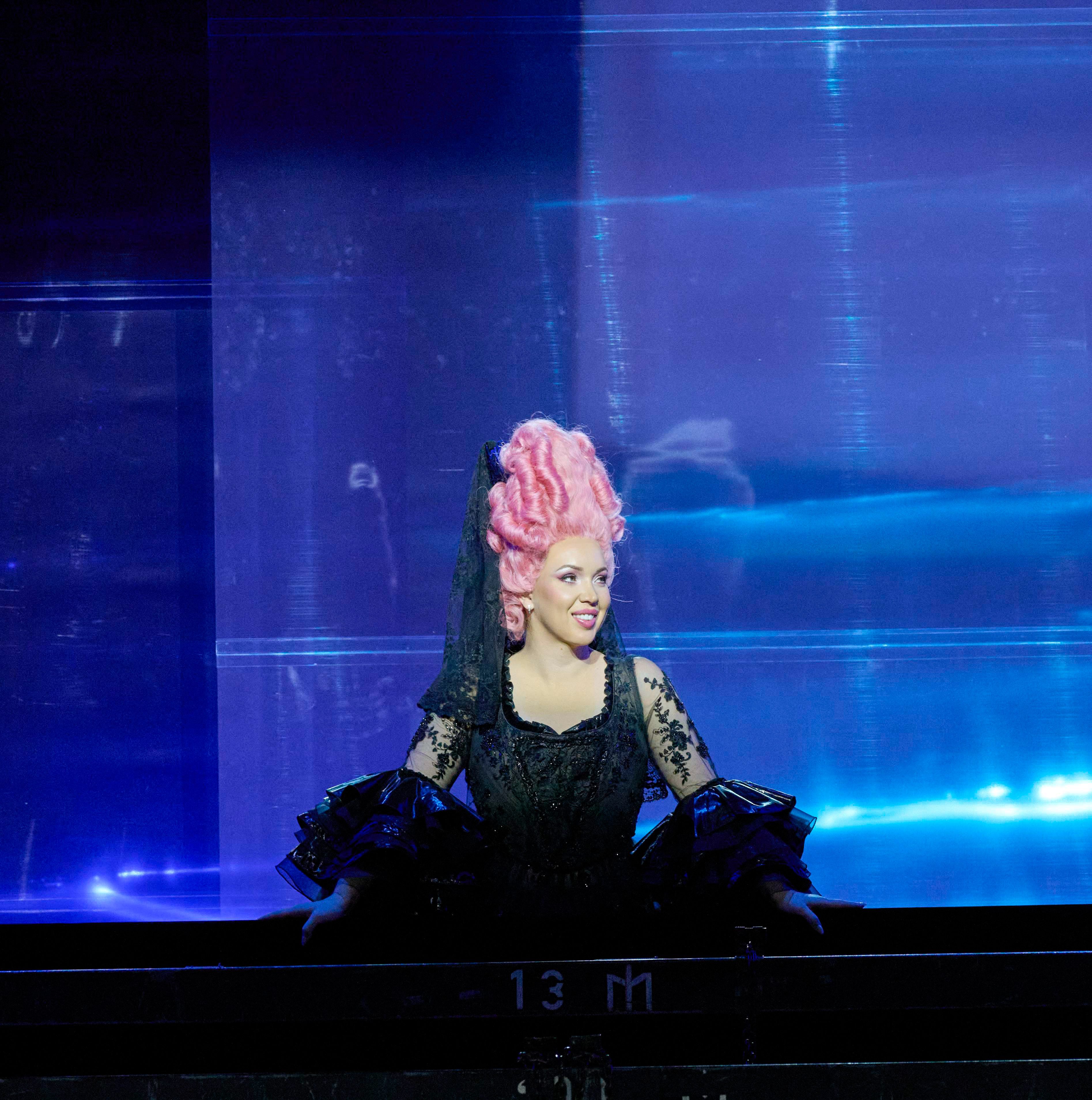 A performer with elaborate pink hair and a black costume sits on stage, smiling. The backdrop features vibrant colors, contributing to a theatrical ambiance. The scene captures a lively moment in a performance, highlighting the character's expressive demeanor.