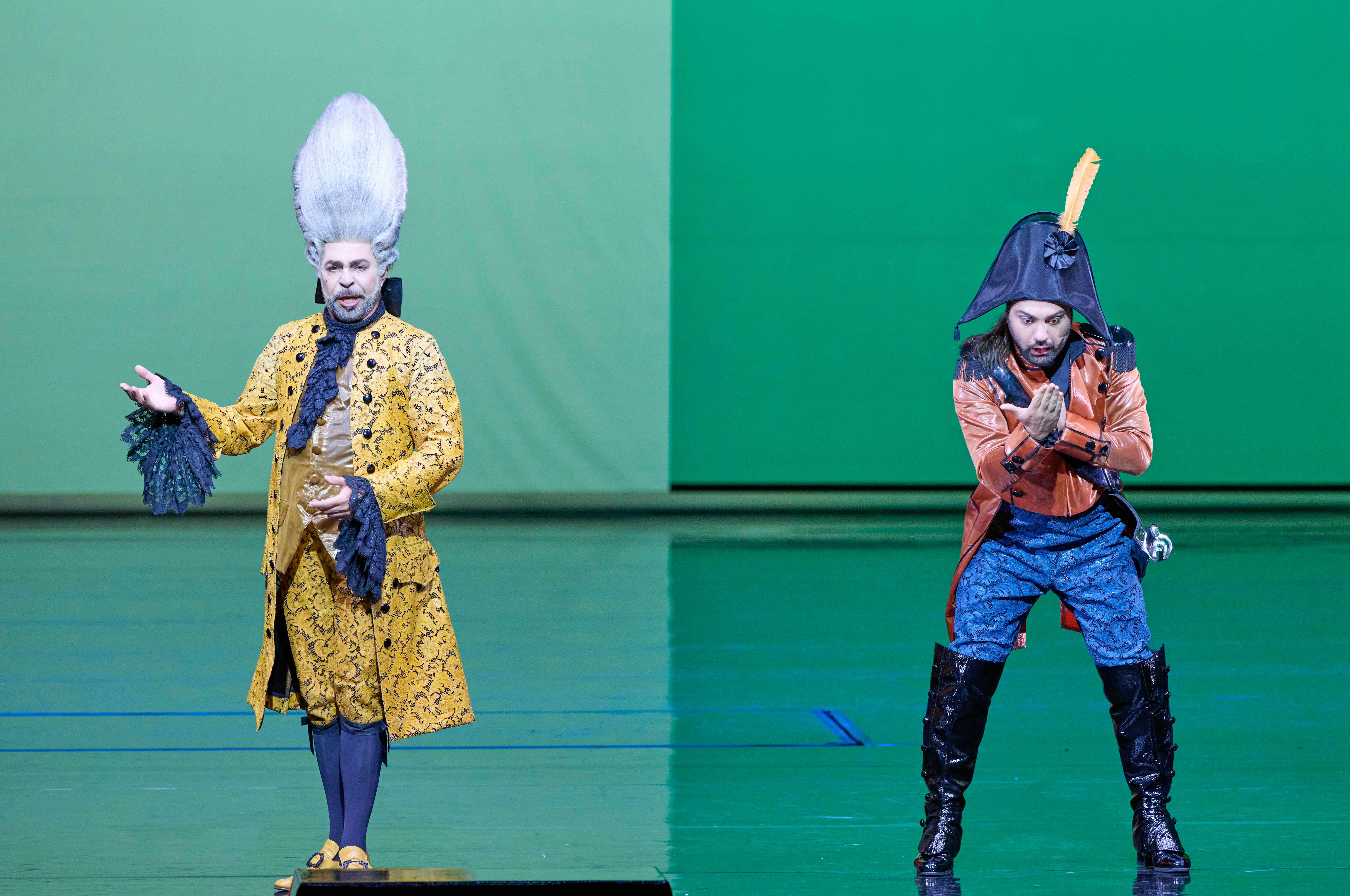 Two male performers stand on stage against a green background. One is dressed in an elaborate yellow costume with a large wig, while the other wears an orange coat and a hat with a feather. They appear engaged in a theatrical performance.