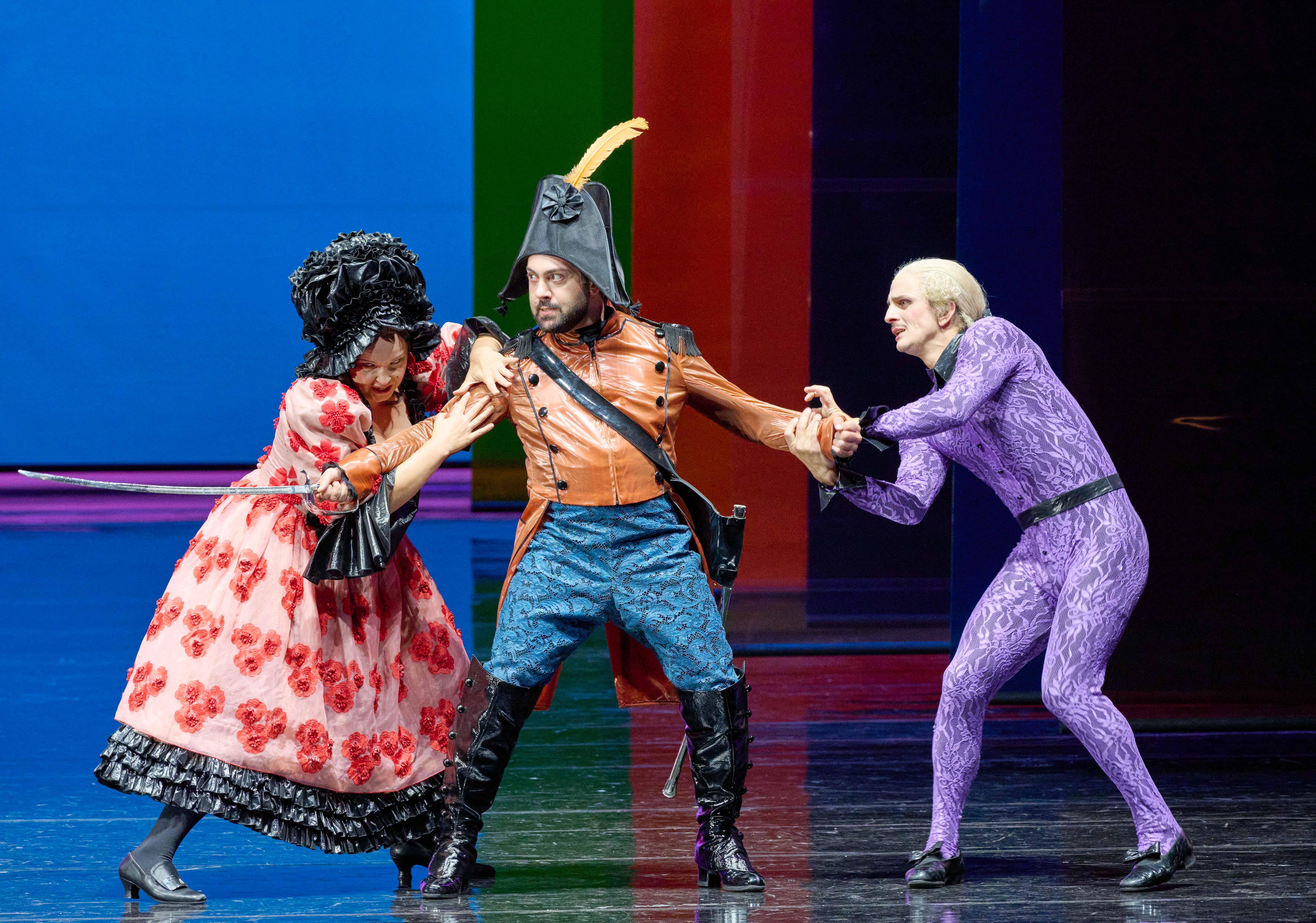 Three performers are engaged in a dramatic confrontation on stage. One wears a vibrant red and black floral dress, holding a sword. The second, dressed in a brown leather jacket and blue pants, stands between them. The third, in a purple outfit, reaches out in a tense scenario.