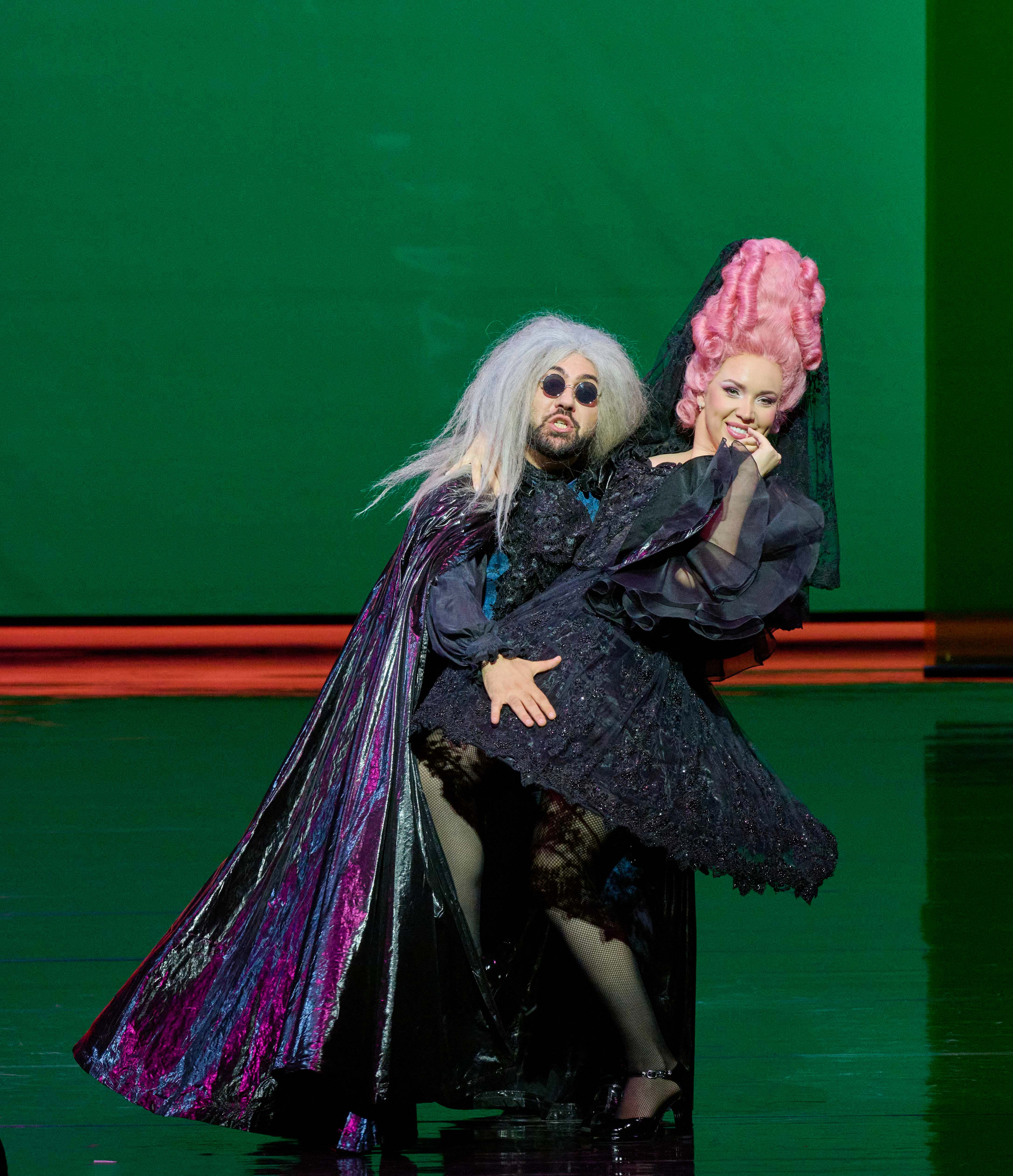 Two performers are on stage, one dressed in an elaborate dark costume with long white hair and sunglasses, while the other has bright pink hair styled in an oversized shape and wears a black dress. They are striking a dramatic pose against a vivid green background.