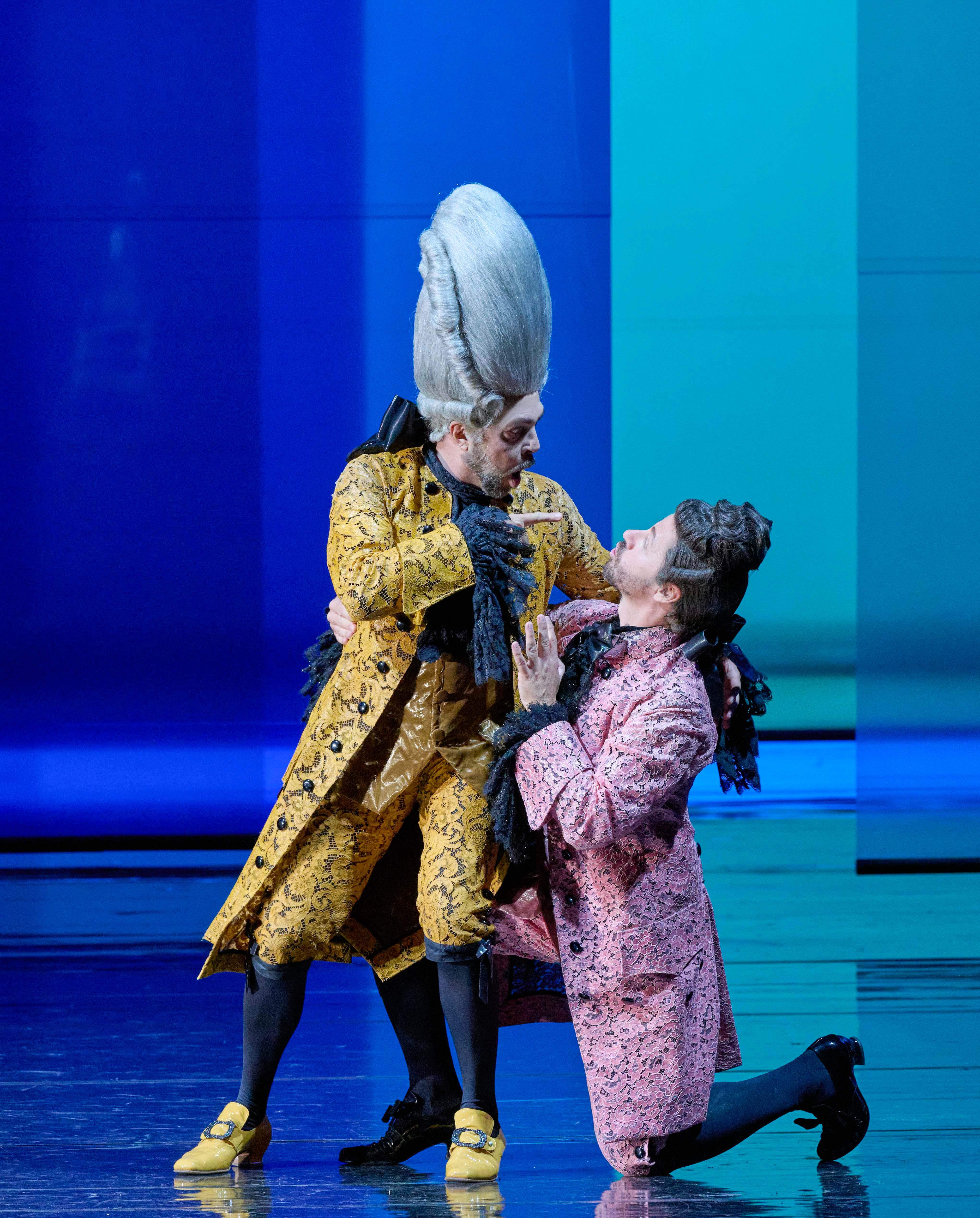 Two male performers are engaged in a dramatic scene, wearing elaborate costumes with intricate patterns. One character, in a yellow coat and tall hairstyle, appears to be assertively addressing the other, who is in a pink coat and is kneeling, suggesting a moment of tension or confrontation.