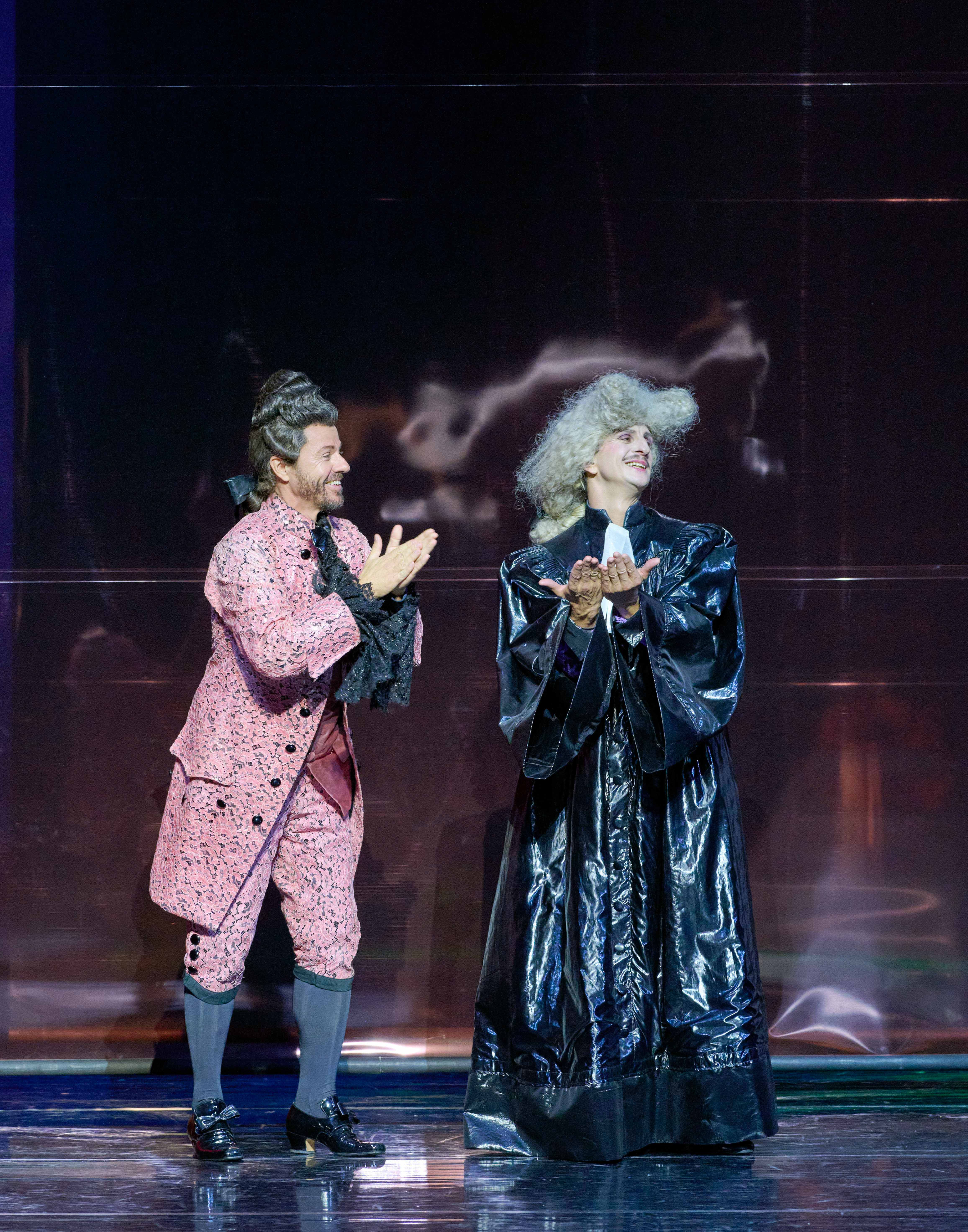 Two performers stand on stage, engaging with each other. One wears a pink, ornate costume and the other is dressed in a long, shiny black robe with curly gray hair. Both are smiling and appear to be in a celebratory moment, applauding or acknowledging an audience.