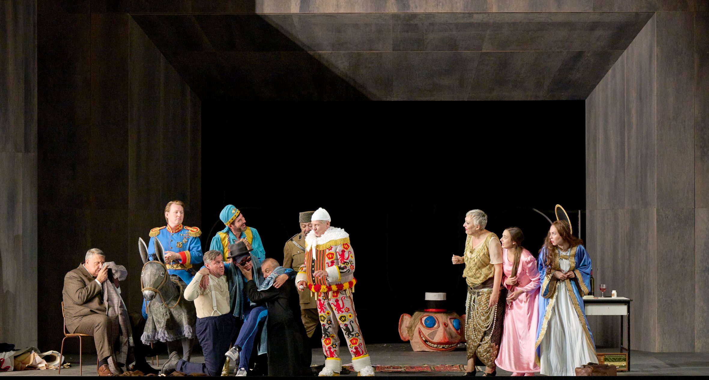 Eine bunt gekleidete Theatergruppe in fantasievollen Kostümen ist auf der Bühne versammelt und wirkt in ein lebhaftes Geschehen vertieft.