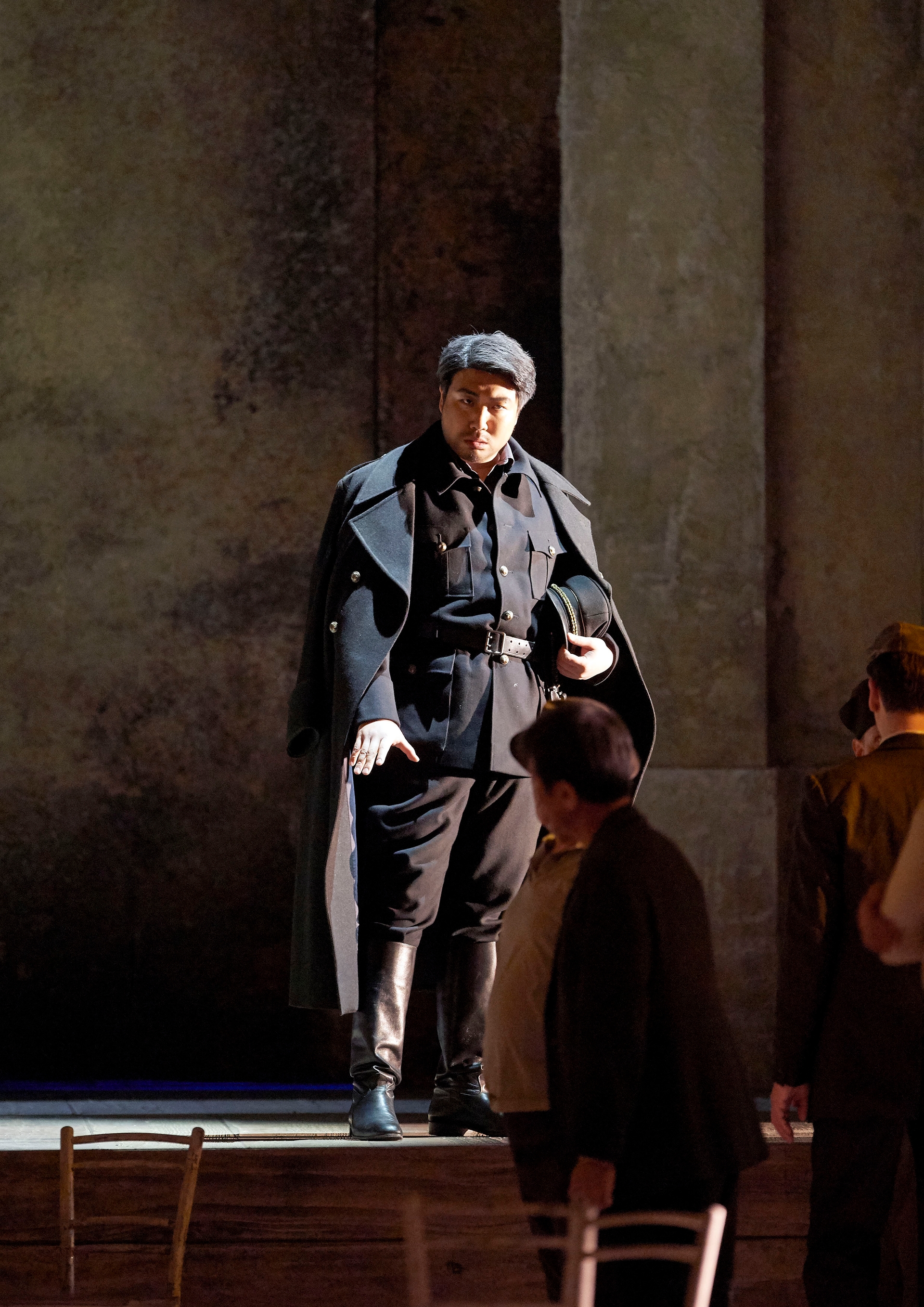 Ein Mann in schwarzer Uniform mit Mantel und Mütze steht streng blickend auf der Bühne, während andere Personen im Hintergrund zu sehen sind.