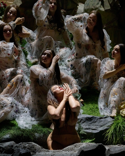 Mehrere Frauen in transparenten, bestickten Gewändern sitzen und knien auf einer grünen Bühne, eine von ihnen hebt emotional die Hände zum Gesicht.