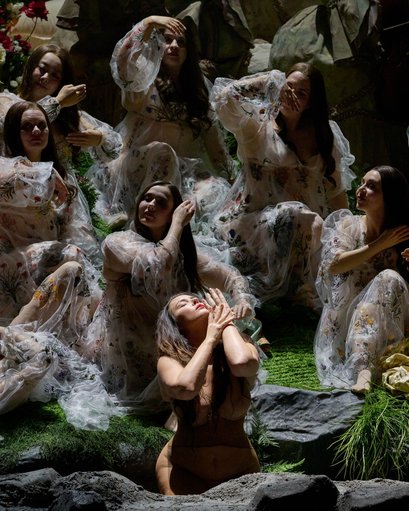 Mehrere Frauen in transparenten, bestickten Gewändern sitzen und knien auf einer grünen Bühne, eine von ihnen hebt emotional die Hände zum Gesicht.