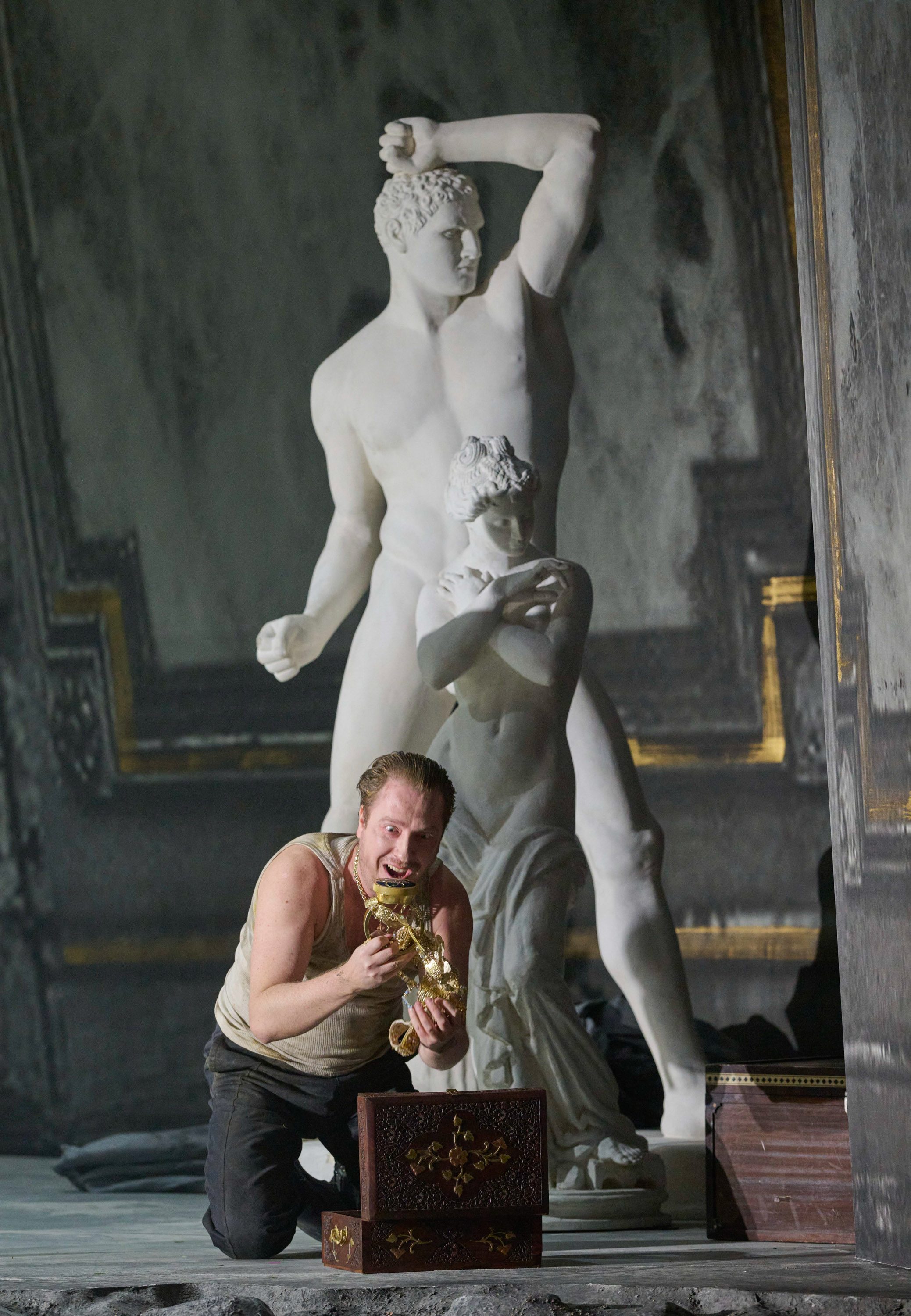 Ein Mann kniet vor einer geöffneten Truhe und betrachtet gierig eine goldene Figur, hinter ihm steht eine große Marmorskulptur.