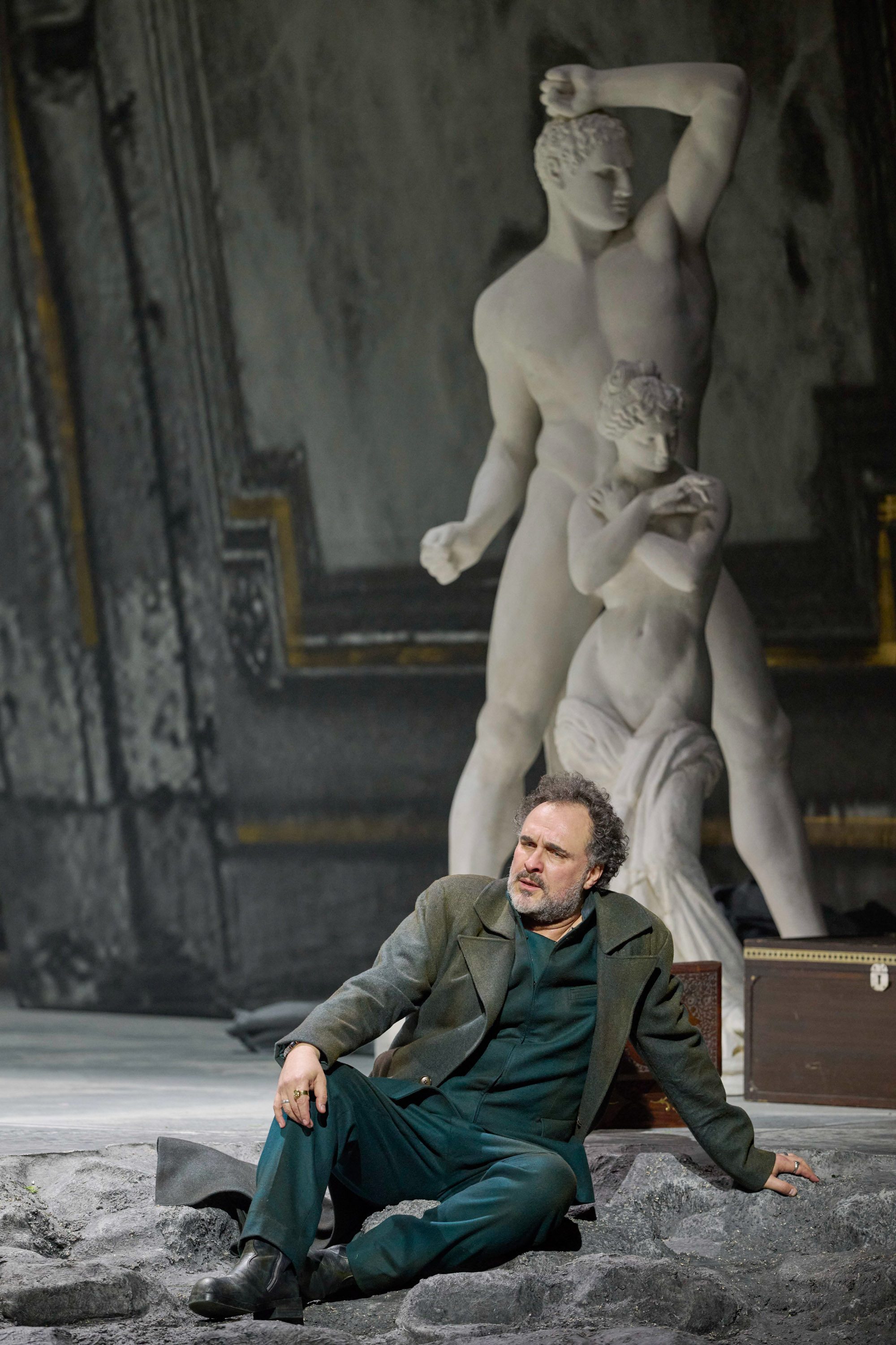 Ein Mann in grünem Anzug sitzt nachdenklich auf steinigem Boden, hinter ihm steht eine weiße Marmorskulptur.