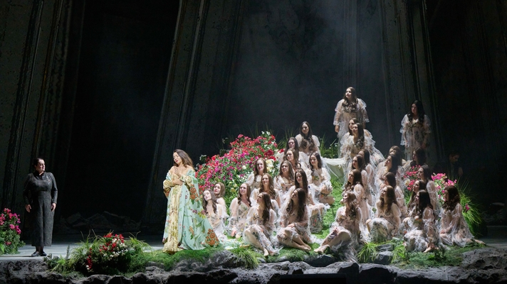 Eine in Gold und Türkis gekleidete Figur steht links auf einer mystischen Blumenbühne, rechts gruppieren sich zahlreiche weiß gekleidete Gestalten auf Terrassen.