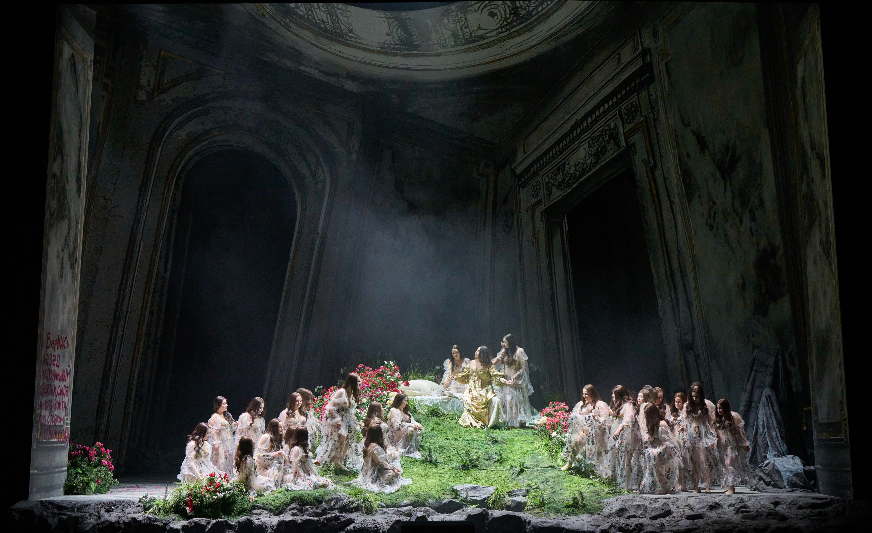 In einer monumentalen Kulisse sitzen und knien zahlreiche Gestalten in weißen Gewändern auf einer grün bewachsenen Bühne, um eine zentral thronende Figur.