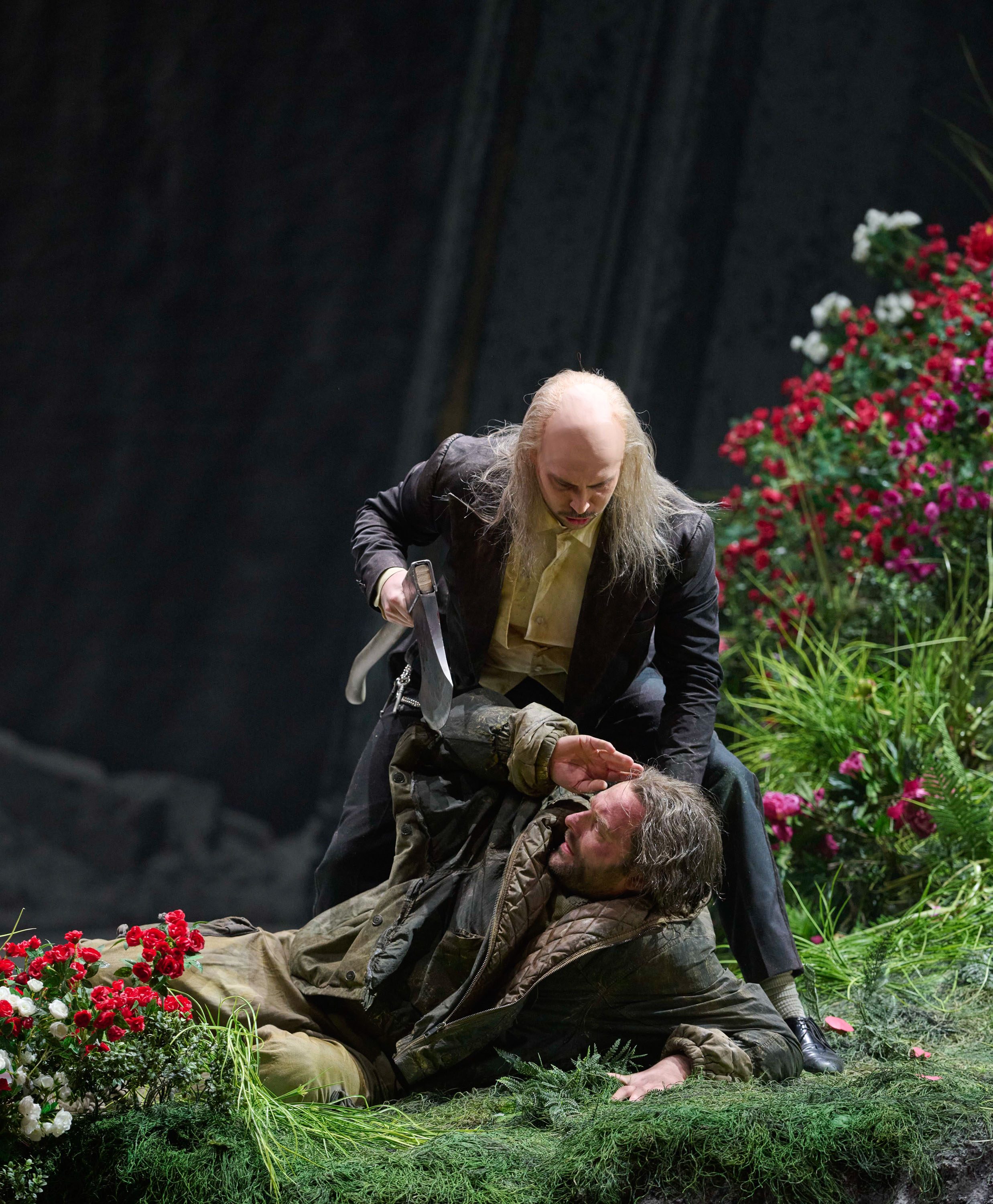 Ein älterer Mann mit Dolch bedroht einen am Boden liegenden Jüngeren auf einer blumenbewachsenen Bühne.